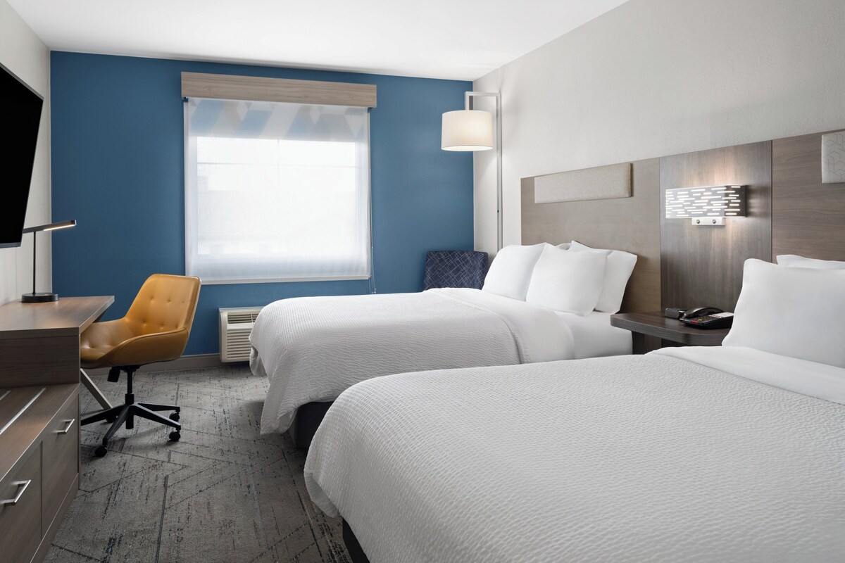 A hotel room with two beds, a desk, a chair, a window with blinds, and a wall-mounted phone.
