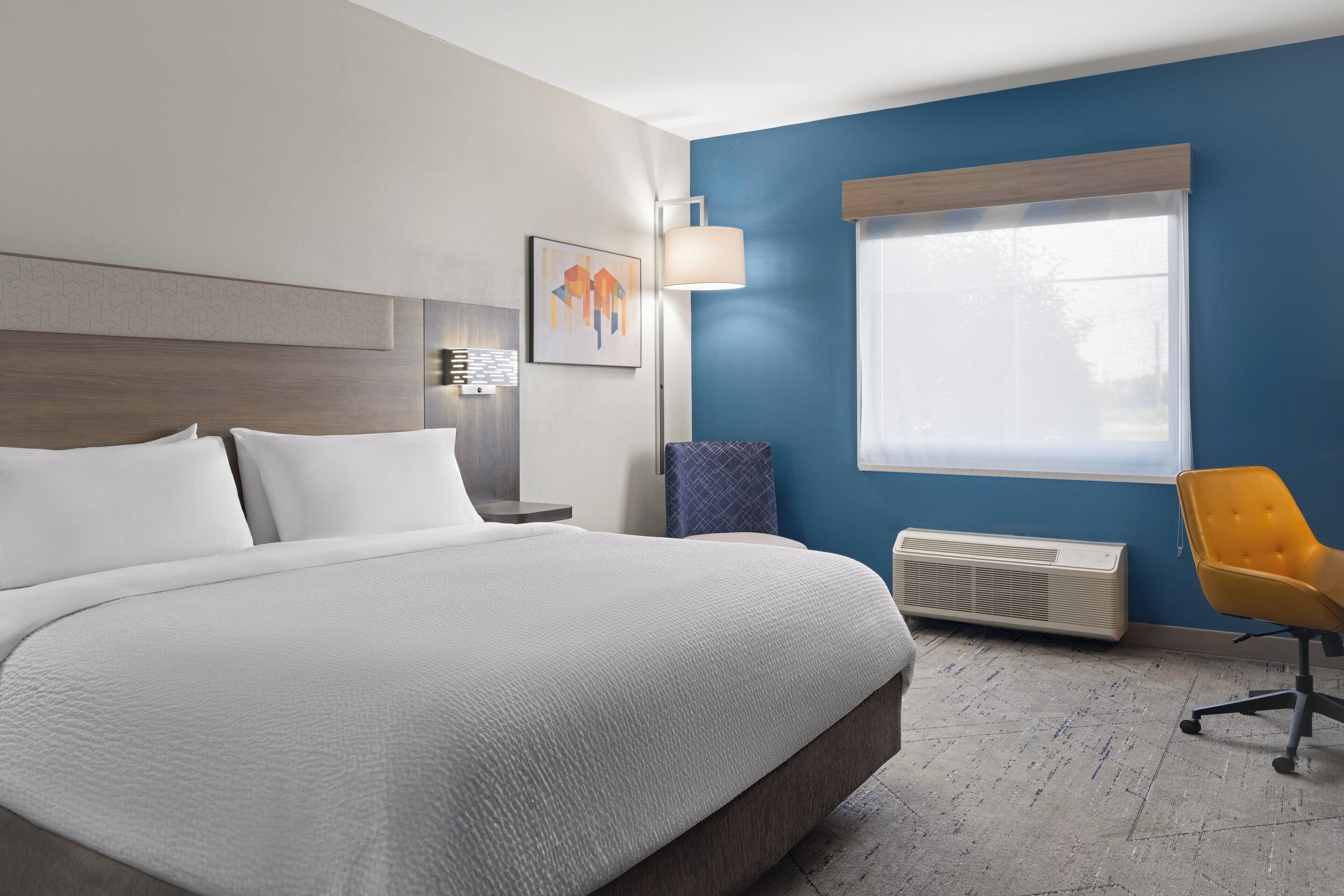 A hotel room with a large bed, a nightstand, a lamp, a chair, and a window with blinds.