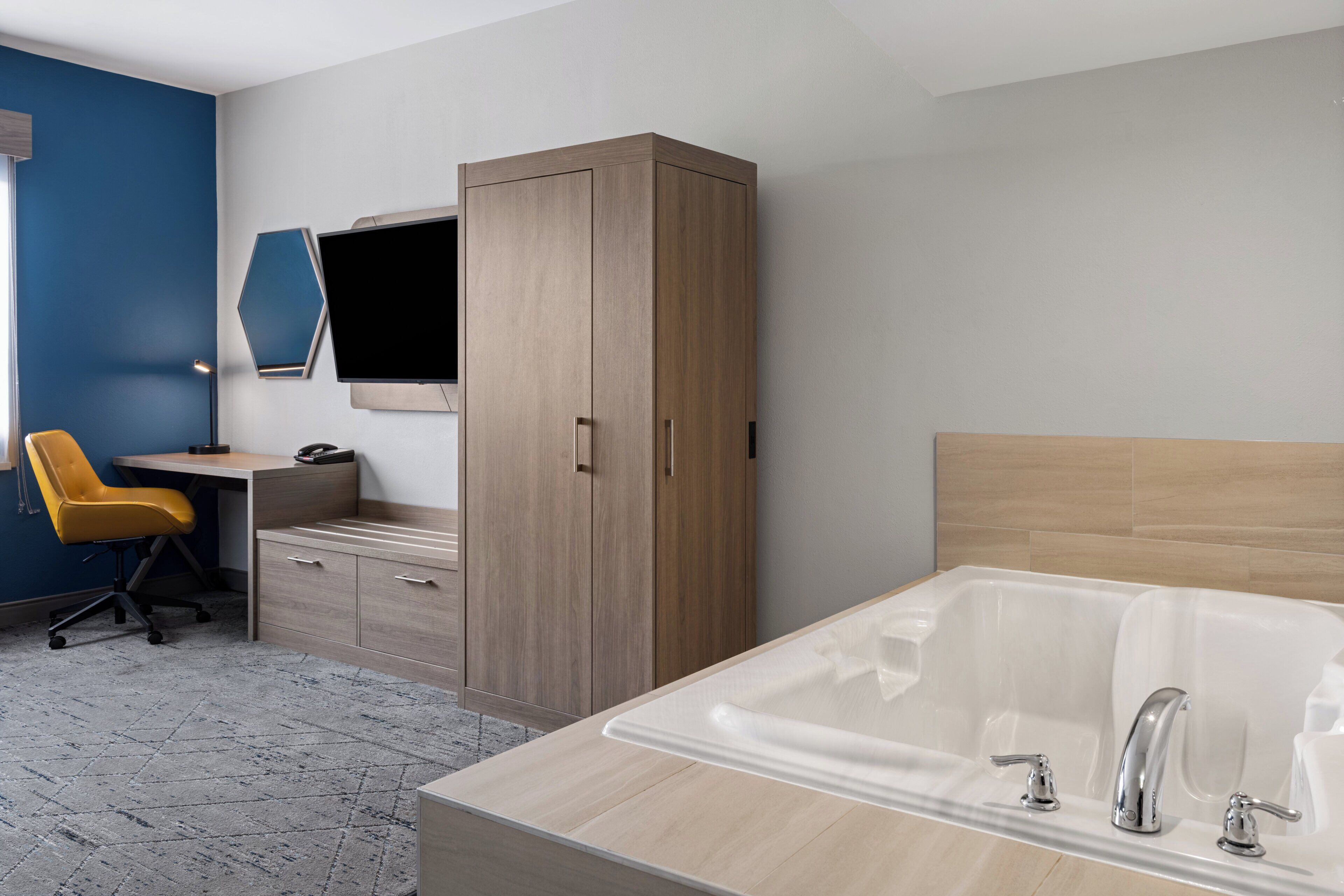 A modern hotel room with a wooden wardrobe, a desk with a chair, a large bathtub, and a flat-screen TV mounted on the wall.
