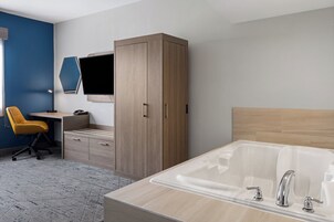 Una habitación de hotel moderna con un armario de madera, un escritorio con una silla, una bañera grande y un televisor de pantalla plana montado en la pared.