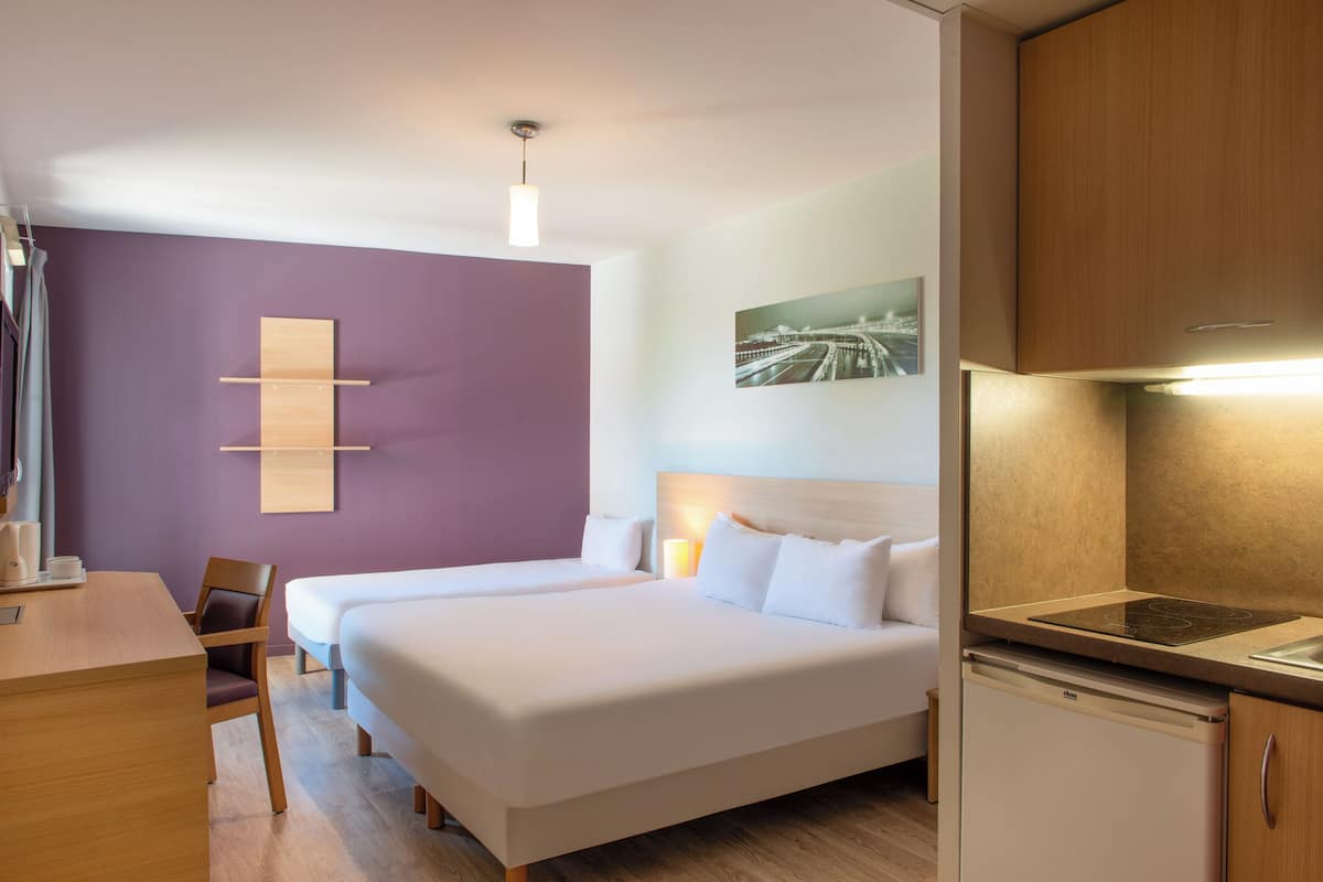 Une chambre d’hôtel avec deux lits, un bureau, une chaise, un réfrigérateur et une étagère fixée au mur.