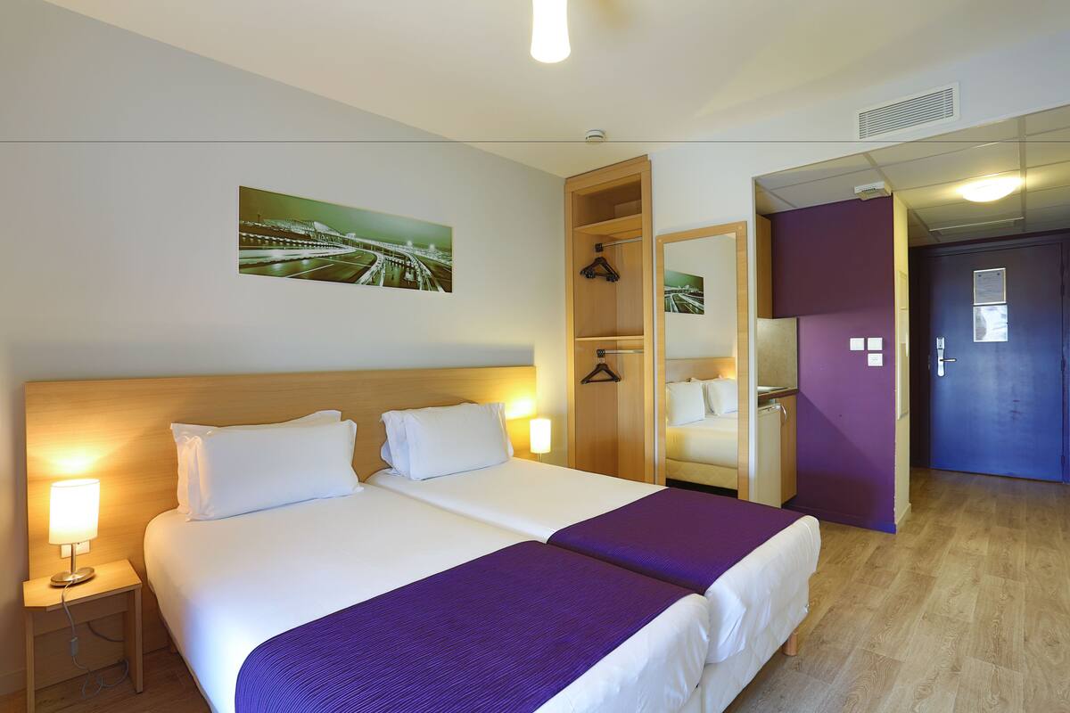 Une chambre d’hôtel avec deux lits, une table de chevet, un miroir et une porte donnant sur une autre pièce.