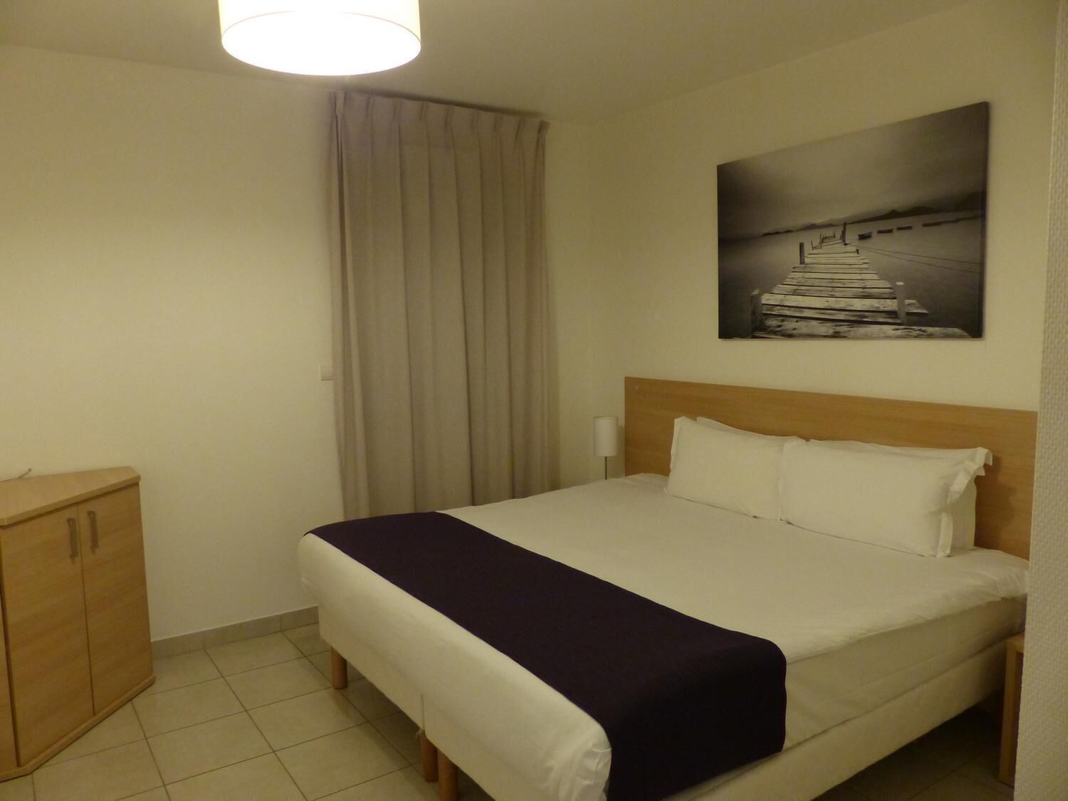 A hotel room with a bed, a wooden cabinet, a painting on the wall, and a window with curtains.