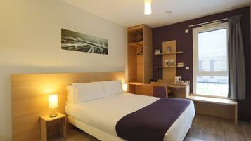 A hotel room with a large bed, bedside table with a lamp, a wooden headboard, a window with curtains, a shelf with items, and a framed picture on the wall.