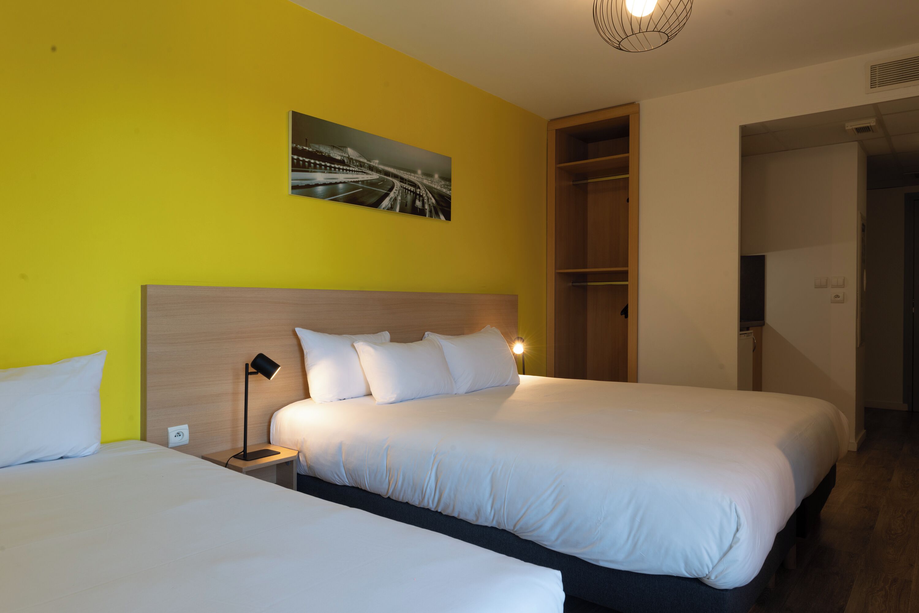Hotelkamer met twee bedden, een houten hoofdbord, een nachtkastje met een lamp, een schilderij aan de muur en een inbouwkast.