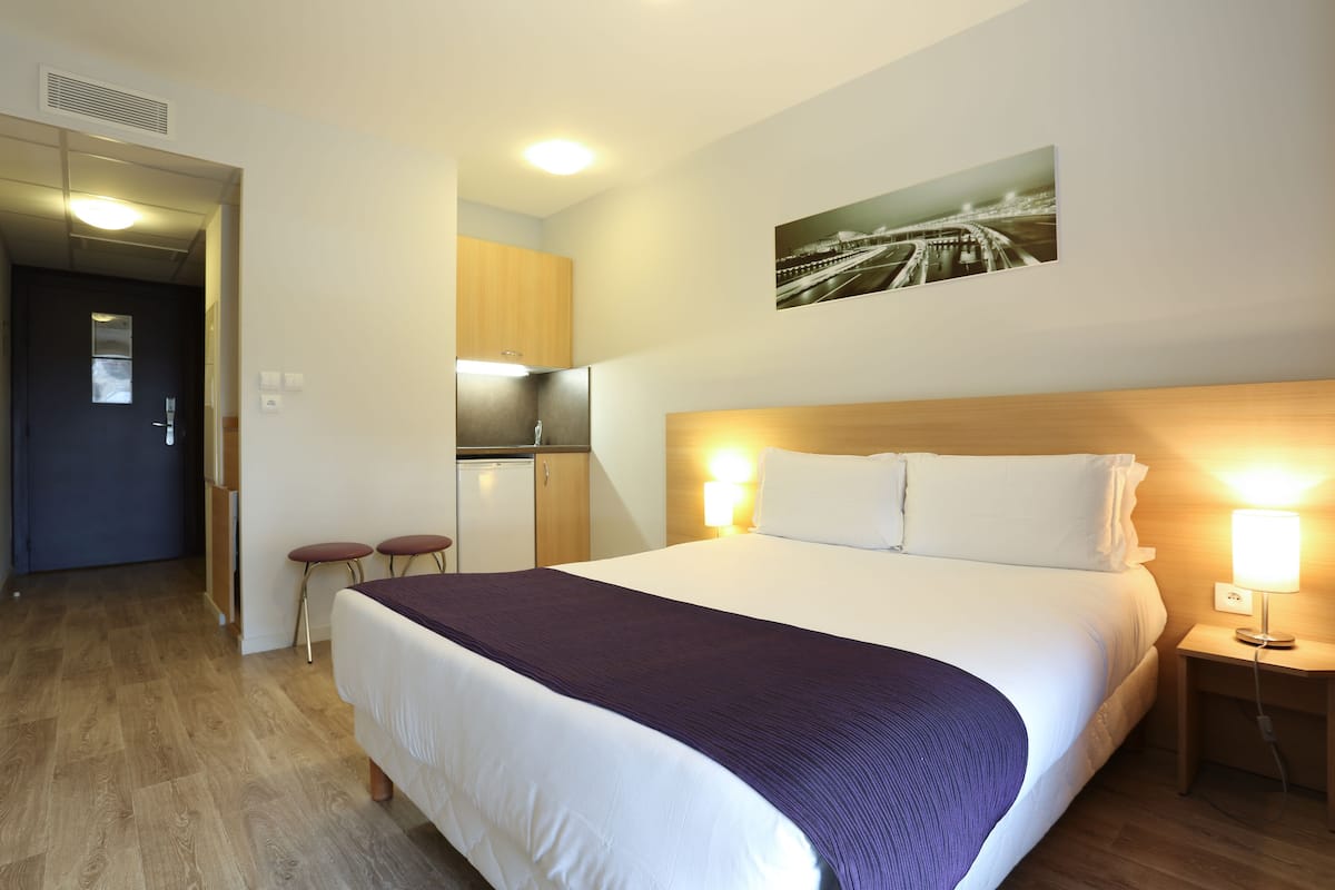 Une chambre d’hôtel avec un grand lit, une tête de lit en bois, une table de chevet avec une lampe, une petite kitchenette et une porte donnant sur une autre pièce.