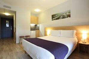 A hotel room with a large bed, a wooden headboard, a nightstand with a lamp, a small kitchenette, and a door leading to another room. - Eastlodge Lyon Est Eurexpo (Saint-Priest)