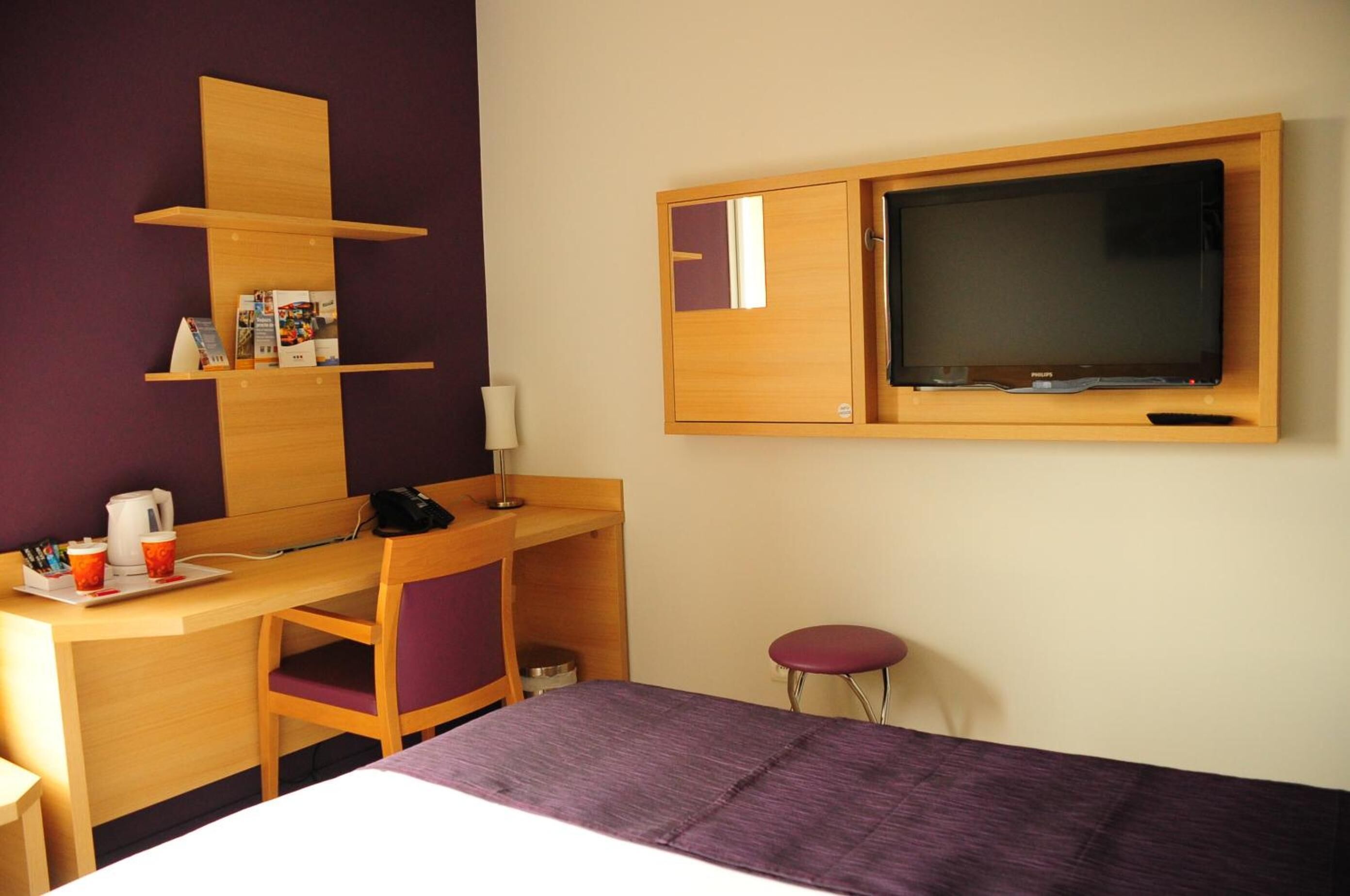 Habitación de hotel con un escritorio de madera, televisor, cama con colcha morada, una silla y un taburete pequeño.