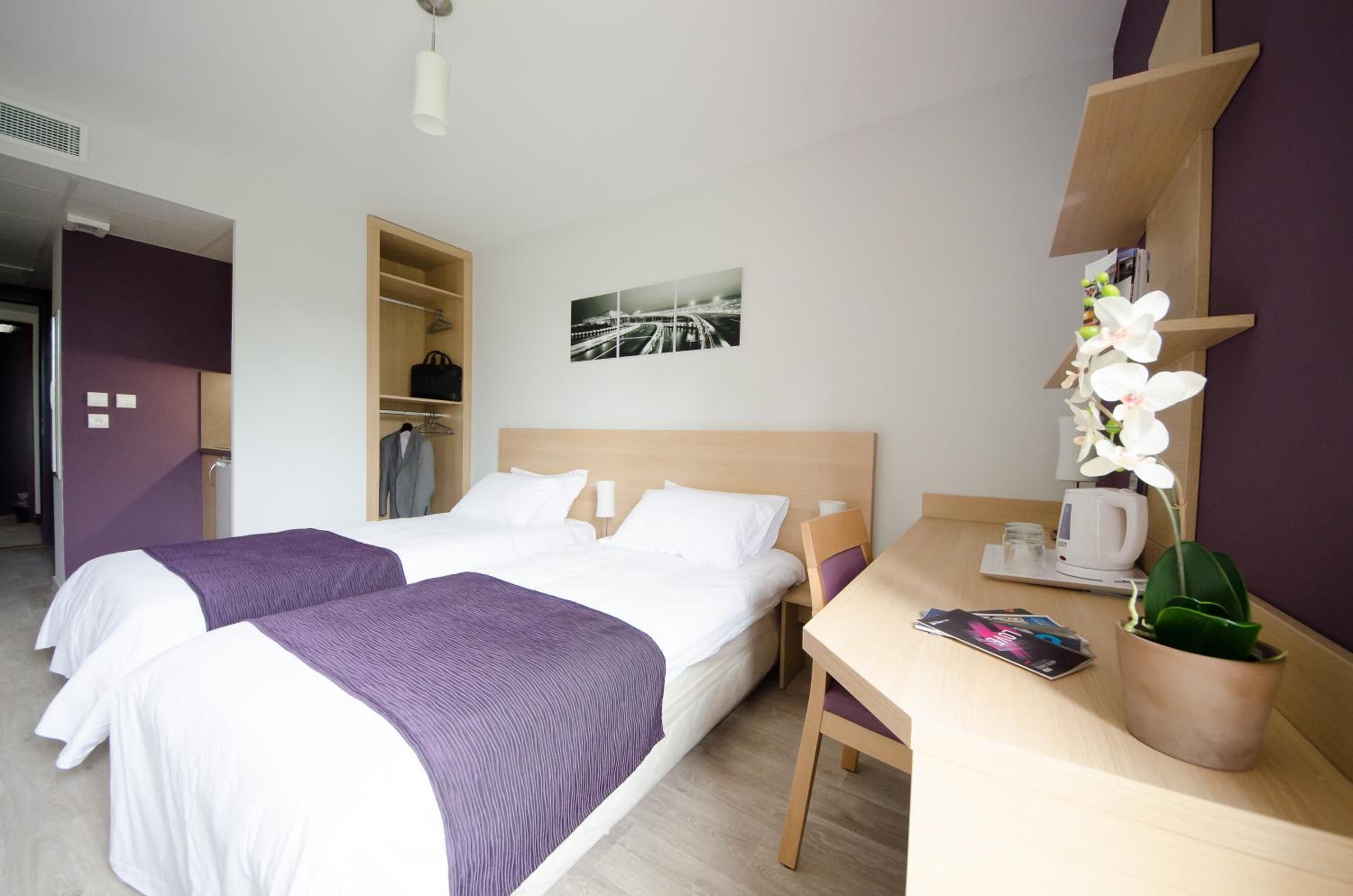 Habitación de hotel con dos camas, un escritorio, una silla, una cafetera, una orquídea en maceta y un cuadro enmarcado en la pared.
