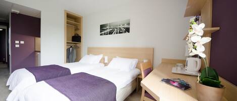 Habitación de hotel con dos camas, un escritorio, una silla, una cafetera, una orquídea en maceta y un cuadro enmarcado en la pared.