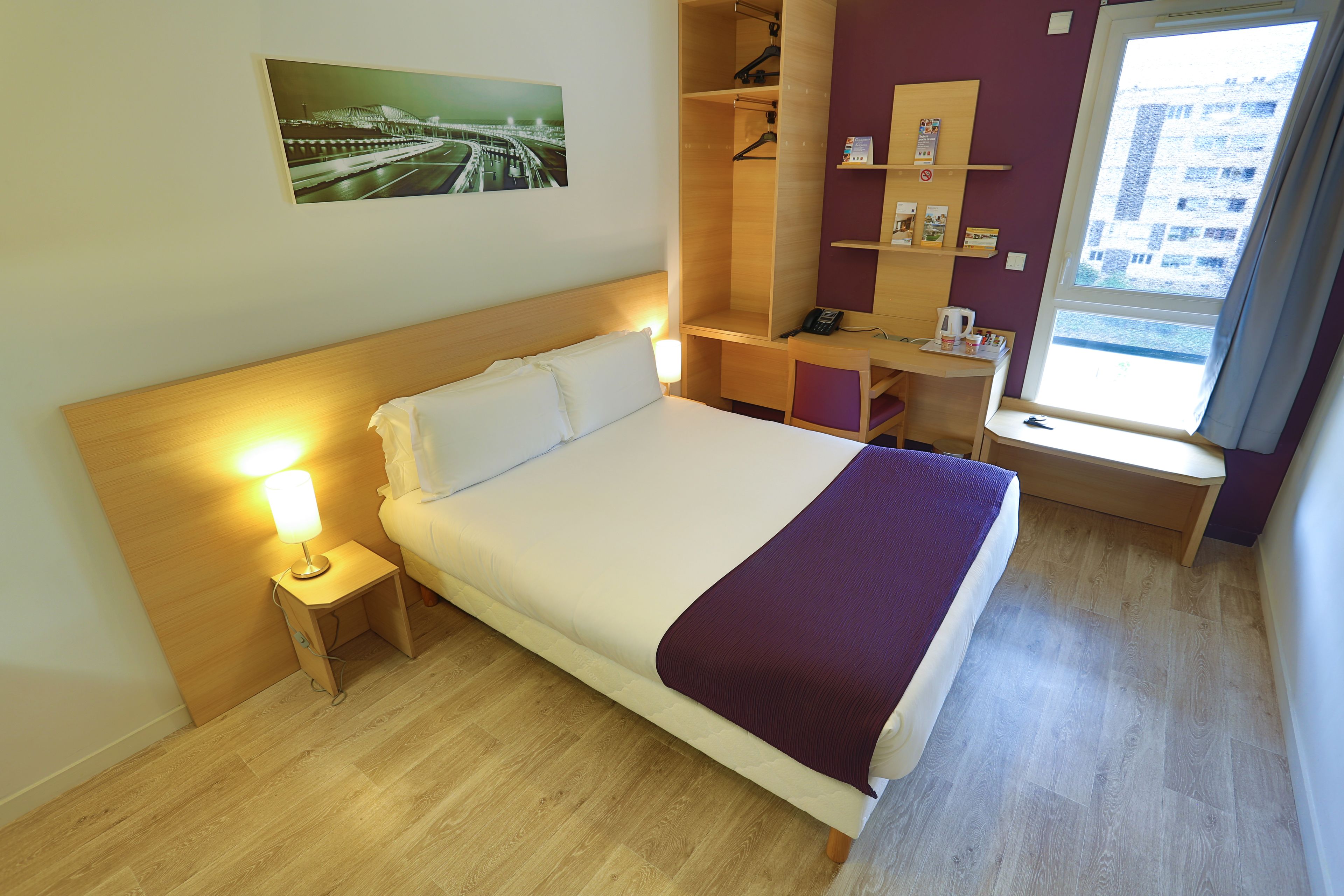 Habitación de hotel con cama, lámpara de noche, suelo de madera, escritorio, silla y ventana con vistas a la ciudad.