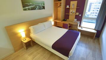Camera d'albergo con un letto, abat-jour, pavimento in legno, scrivania, sedia e una finestra con vista sulla città.