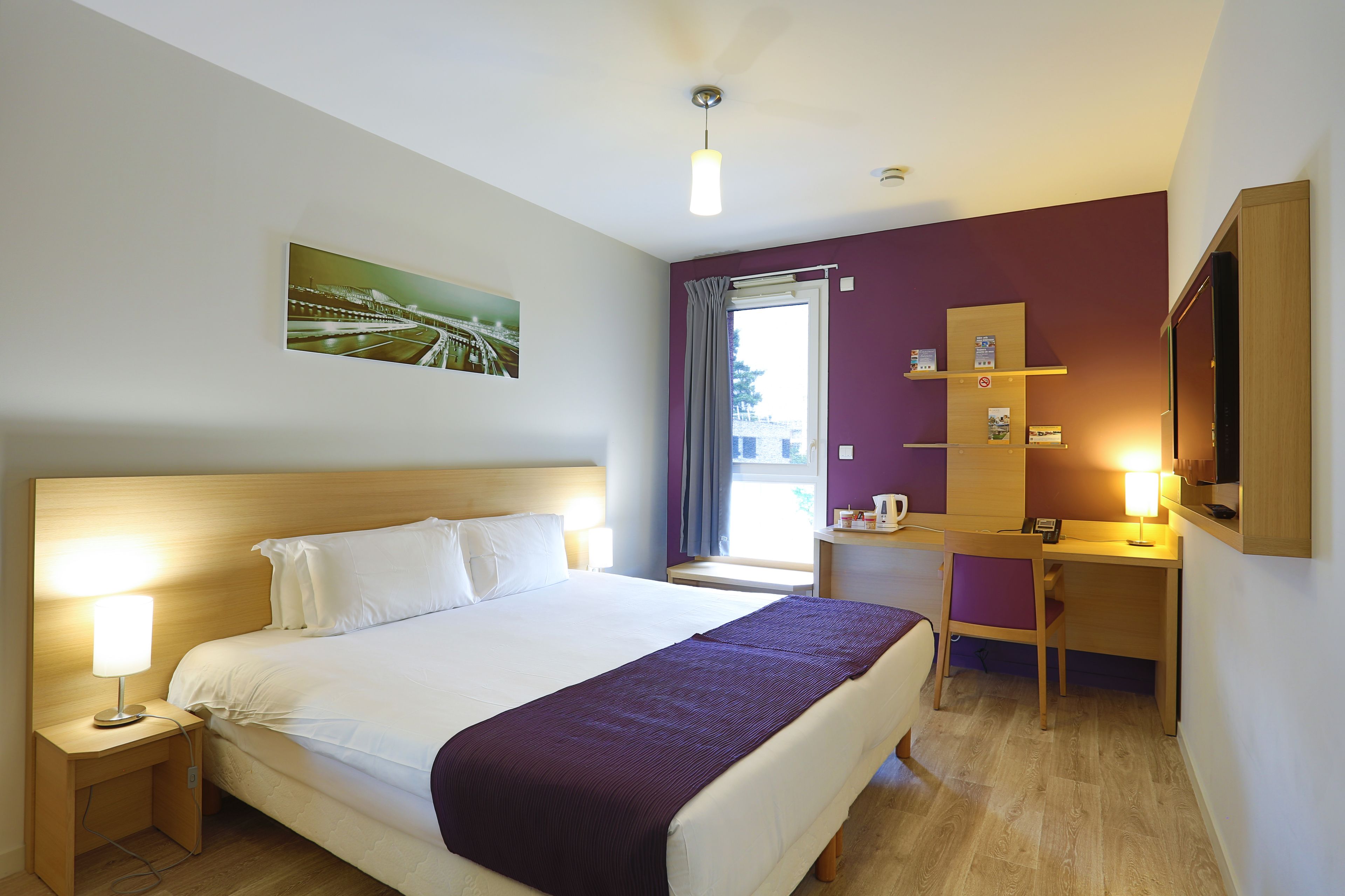 Ein Hotelzimmer mit Bett, Nachttisch, Lampe, Schreibtisch, Stuhl und einem Fenster mit Vorhängen.