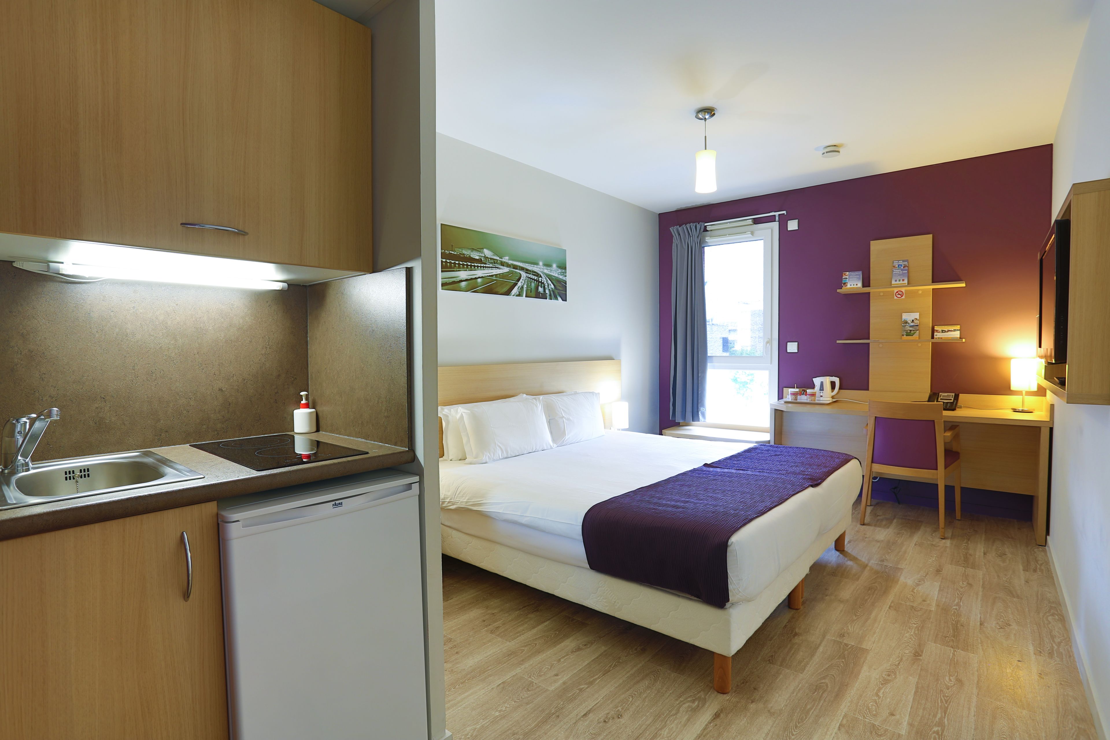 Habitación de hotel pequeña con kitchenette, una cama con manta morada, un escritorio y una ventana con cortinas.