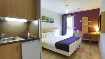 Een compacte hotelkamer met kitchenette, een bed met paars dekbed, een bureau en een raam met gordijnen.