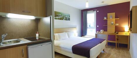Habitación de hotel pequeña con kitchenette, una cama con manta morada, un escritorio y una ventana con cortinas.
