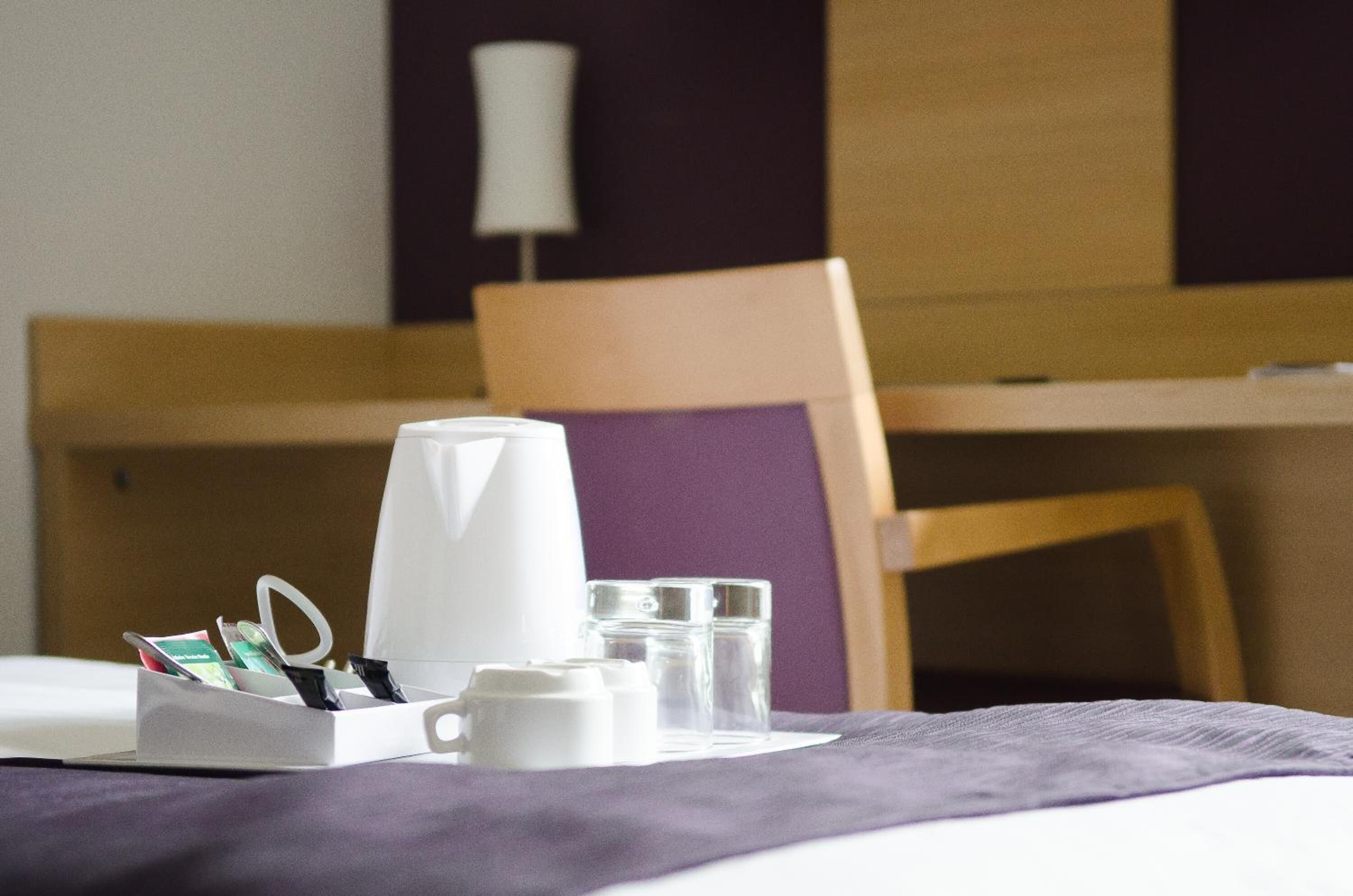 A neatly arranged hotel room with a bed, a bedside table with a coffee maker, glasses, and a tissue box.