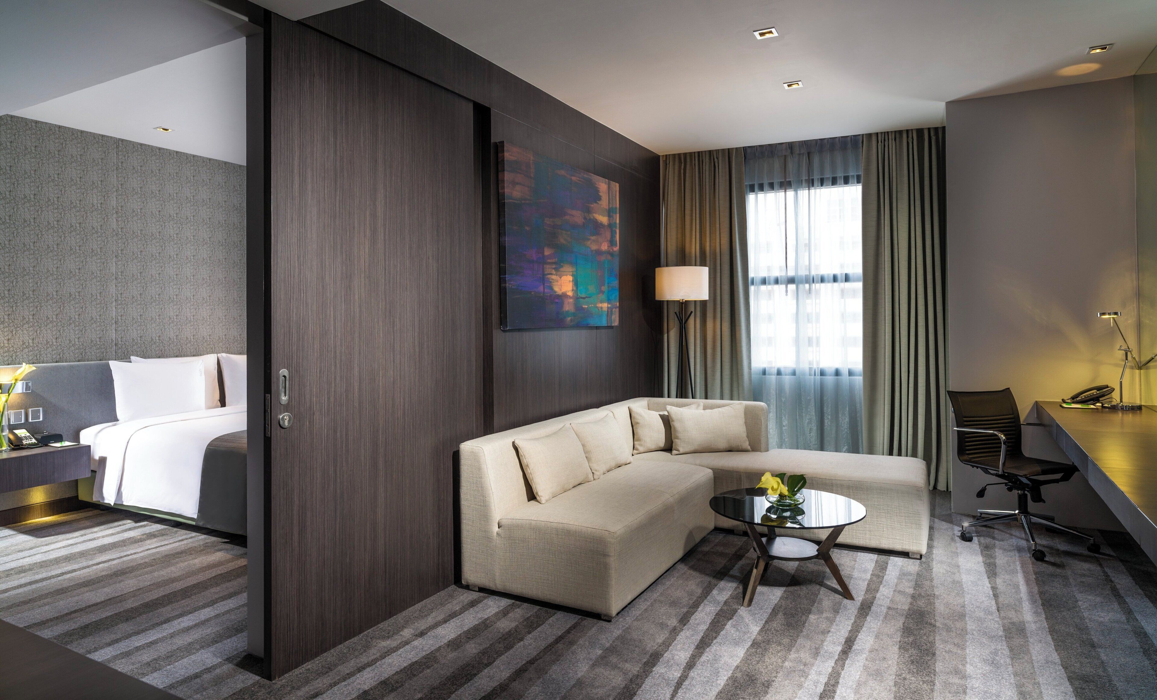 A modern hotel room with a sofa, a small table, a desk with a lamp, and a painting on the wall.