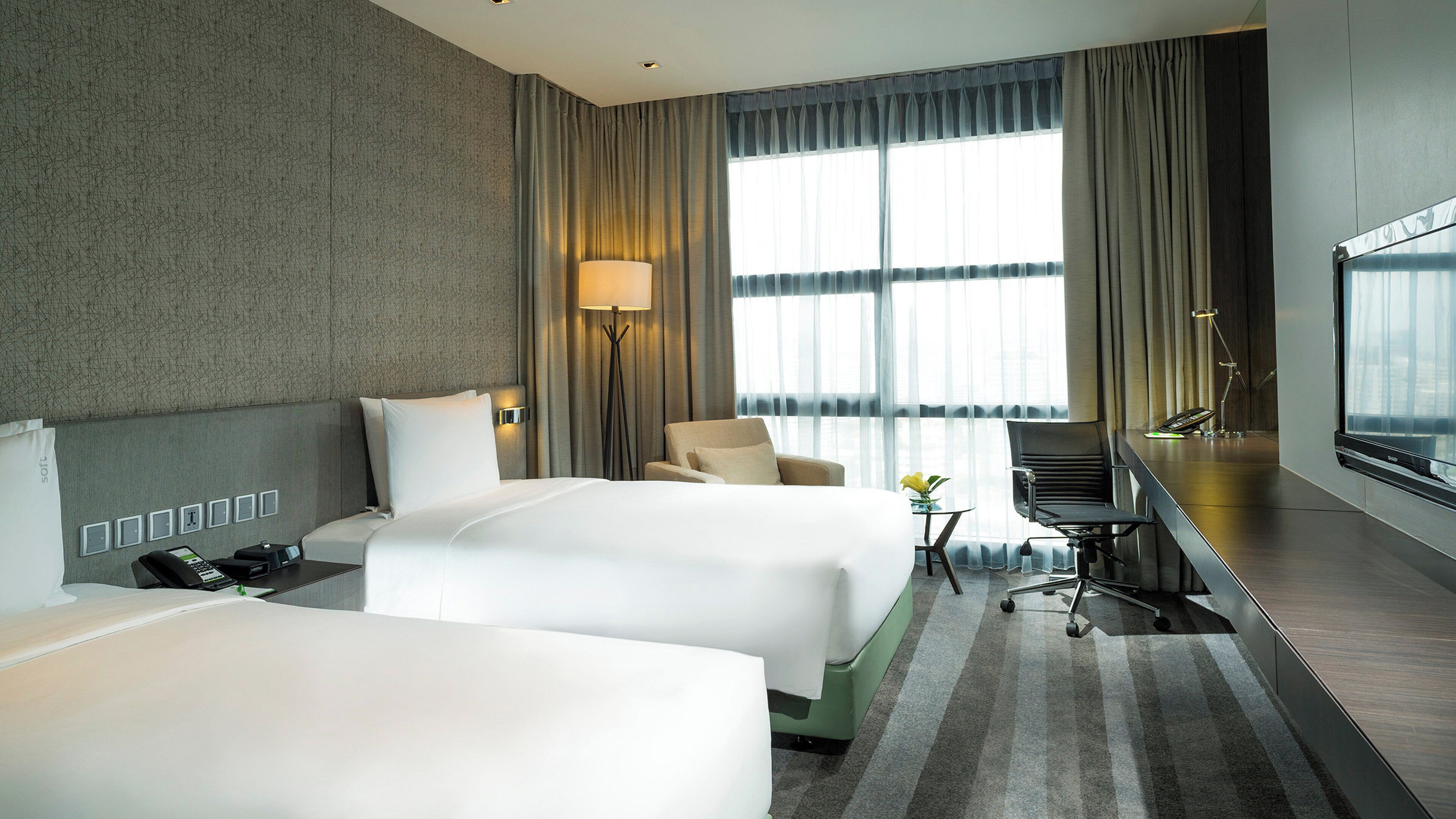 A hotel room with two beds, a desk, a chair, a lamp, and a large window.