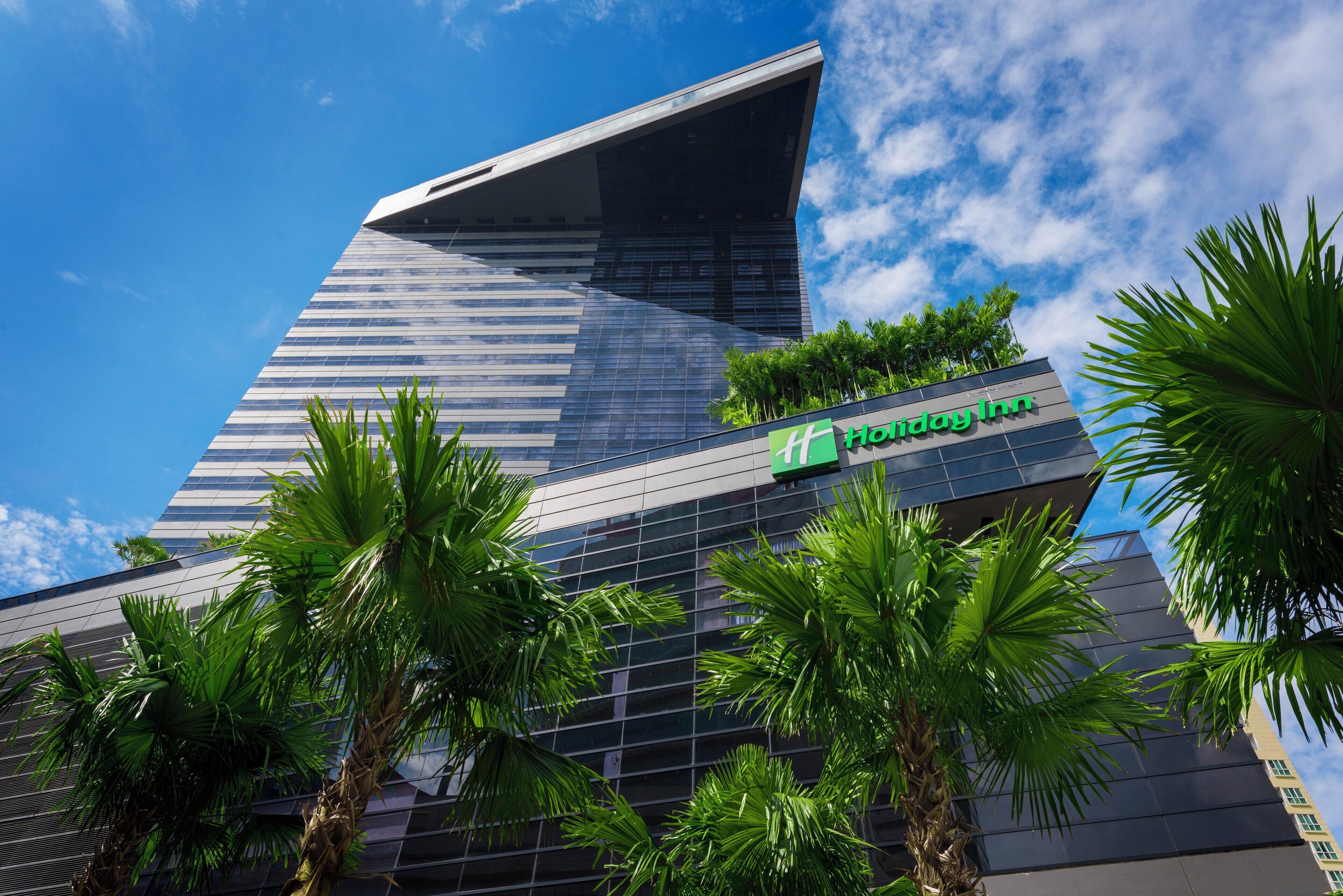 A modern hotel building with a prominent glass facade, a visible logo, and surrounding palm trees.