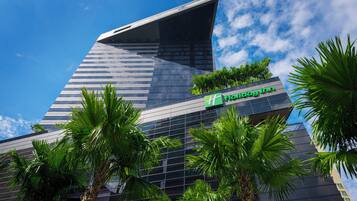 A modern hotel building with a prominent glass facade, a visible logo, and surrounding palm trees.