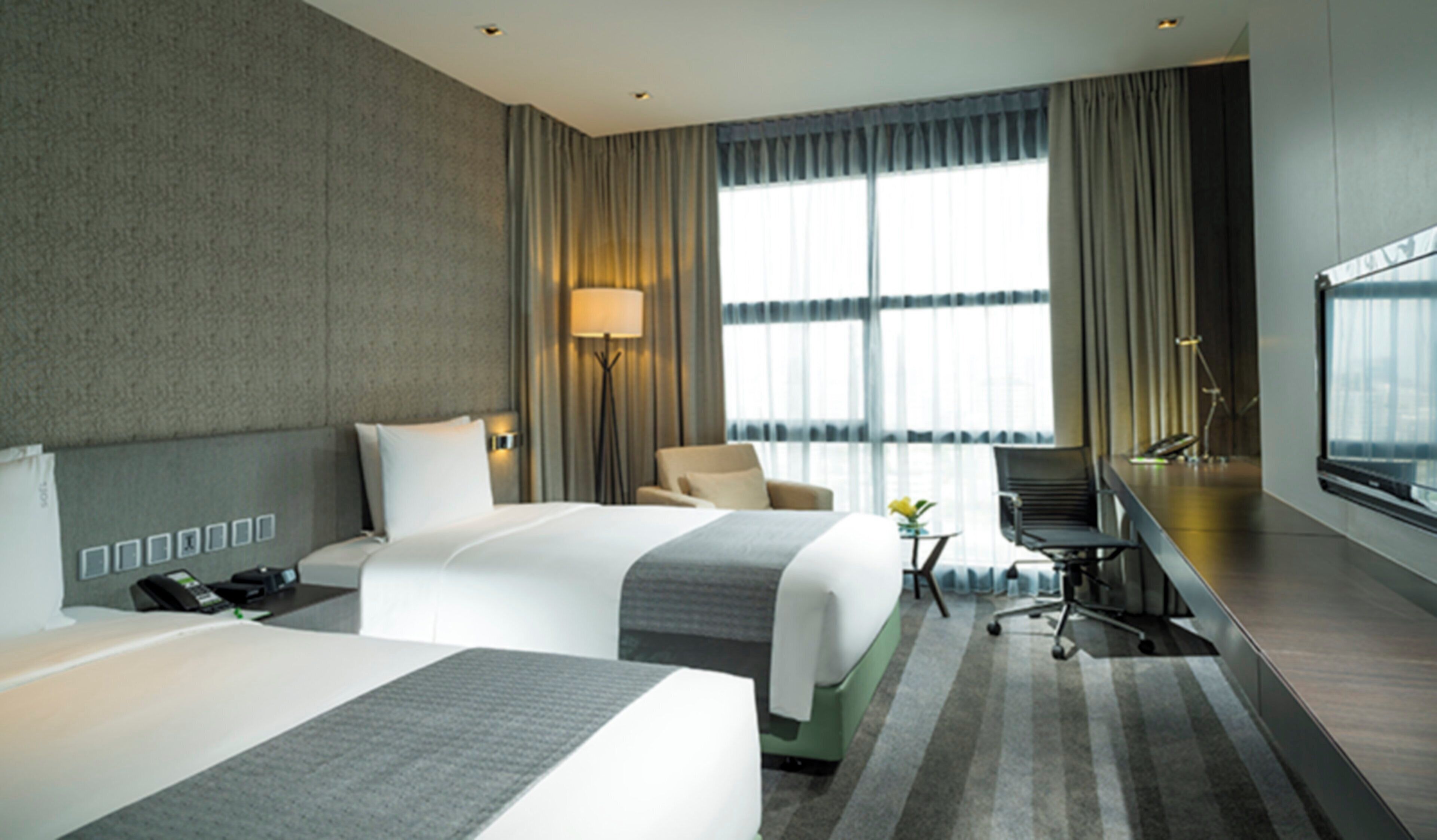 Une chambre d’hôtel avec deux lits, un bureau, une chaise, une lampe et une grande fenêtre avec des rideaux.