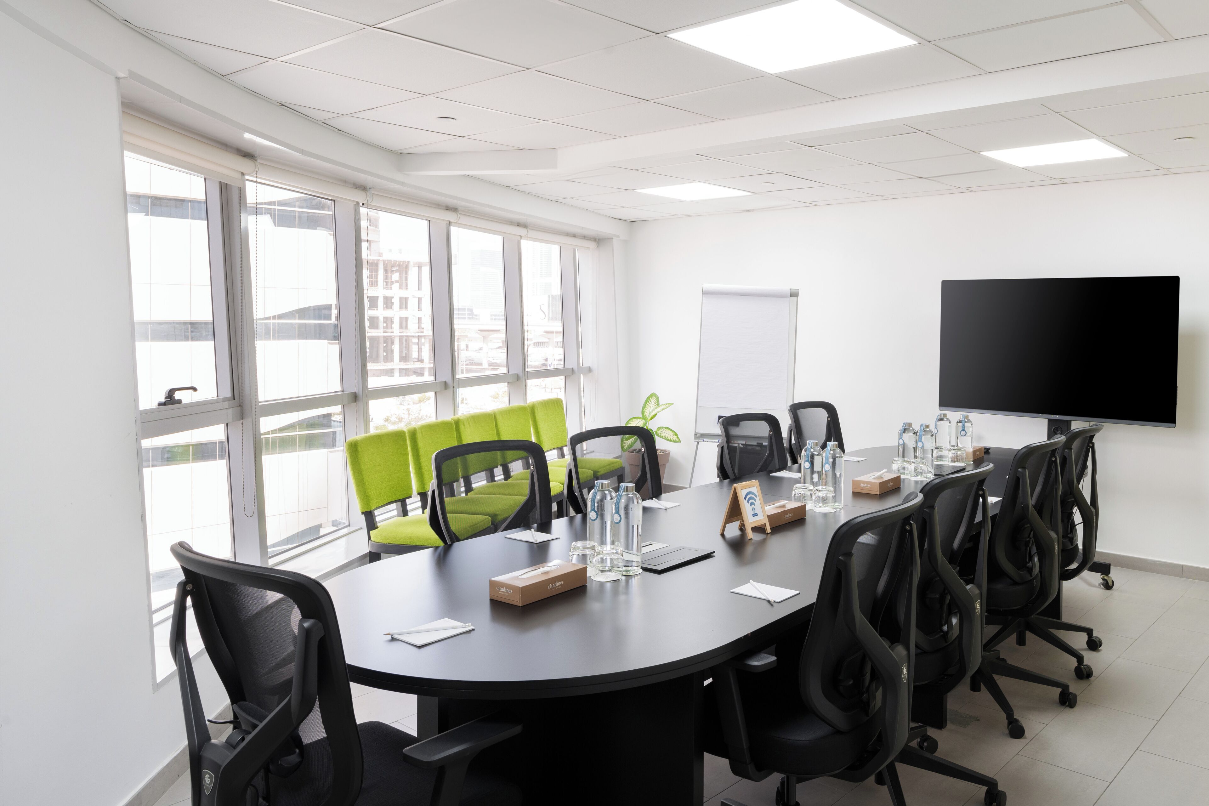 A conference room with a large table, black office chairs, a flat-screen TV, and a window with a view of buildings.