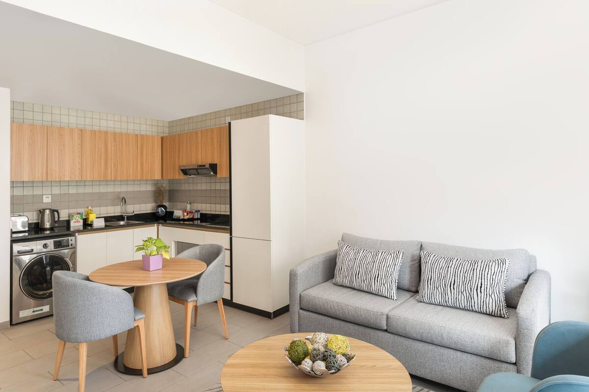 A modern kitchen with wooden cabinets, a dining table with chairs, and a grey sofa with patterned cushions.