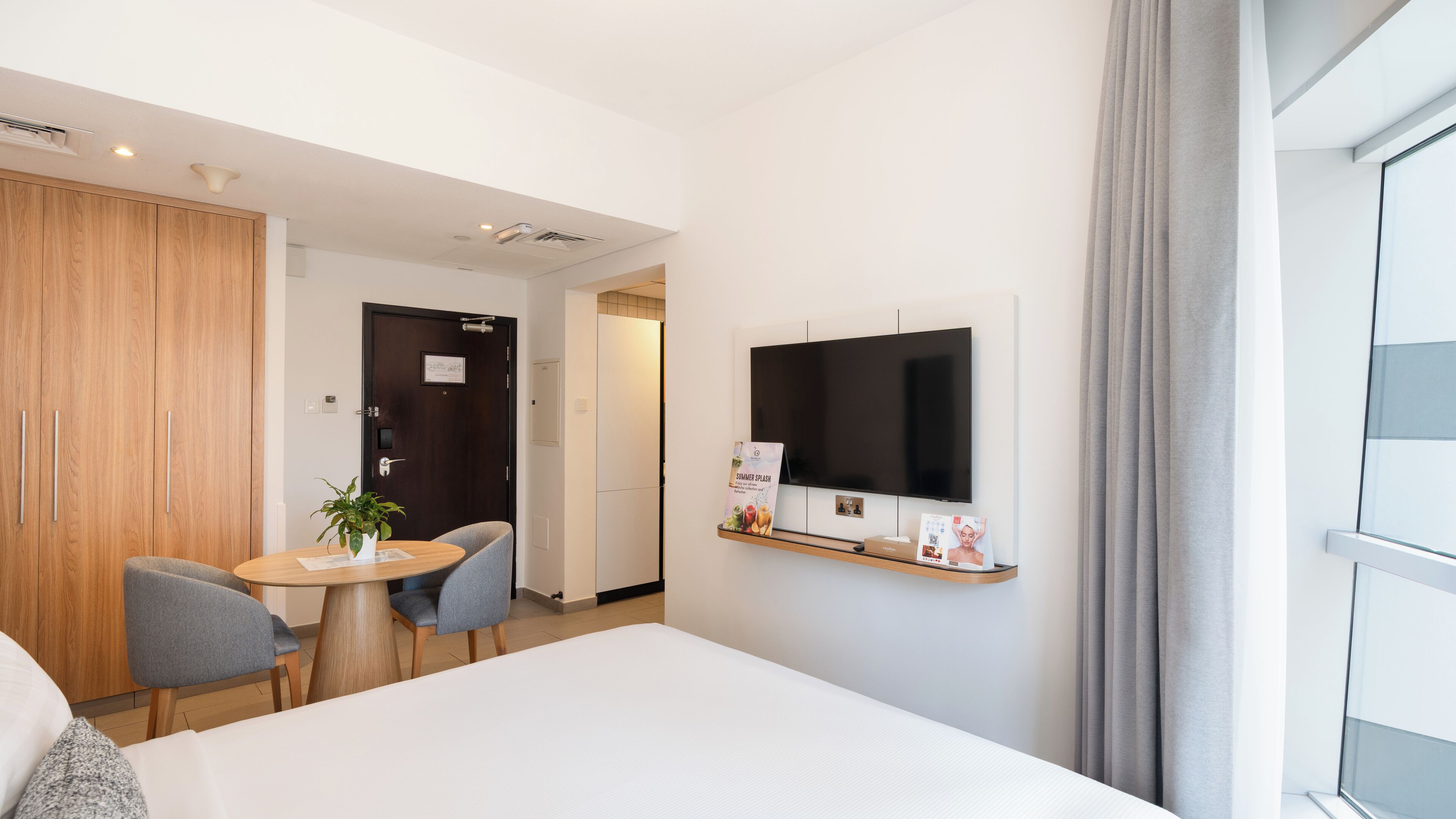 Une chambre d’hôtel avec un lit, un téléviseur fixé au mur, une petite table avec des chaises, une armoire en bois et une plante en pot.