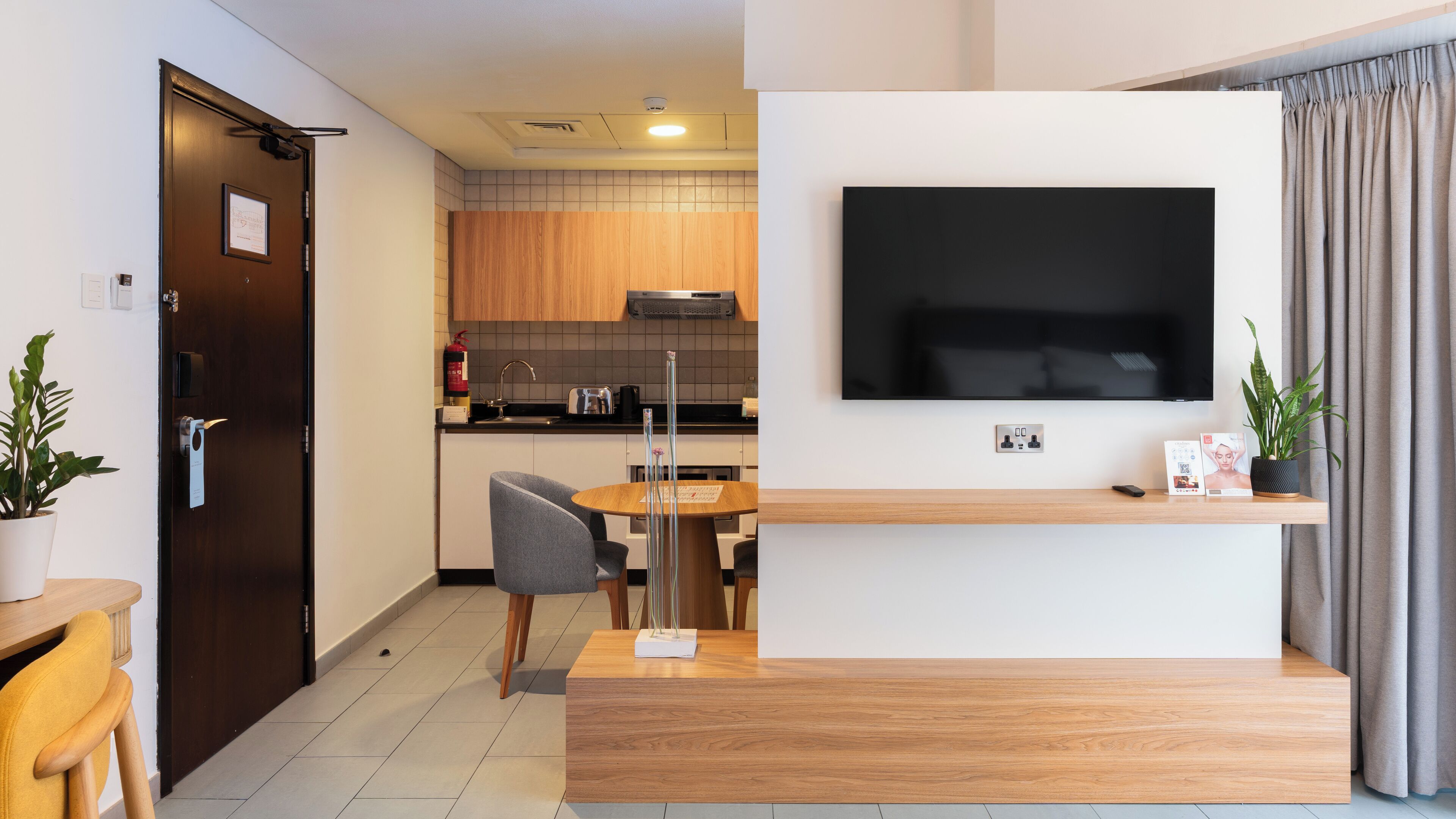 Ein modernes Wohnzimmer mit Flachbildfernseher an der Wand, einem Holztisch, einem Essbereich mit kleinem Tisch und Stühlen sowie einer Küchenzeile mit Holzschränken und einer Mikrowelle.