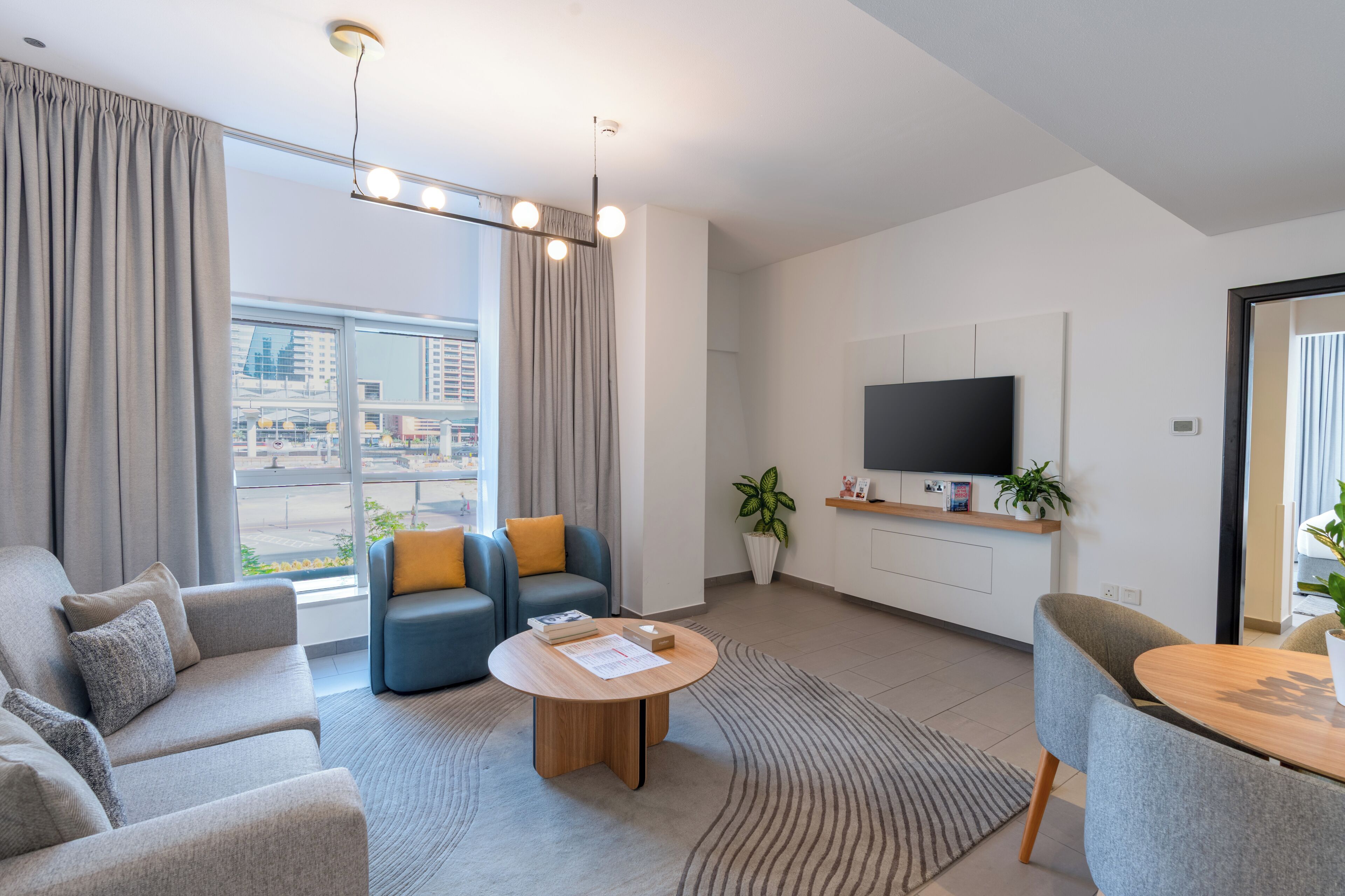 A modern living room with a sofa, armchairs, a coffee table, a flat-screen TV, and a large window with curtains.