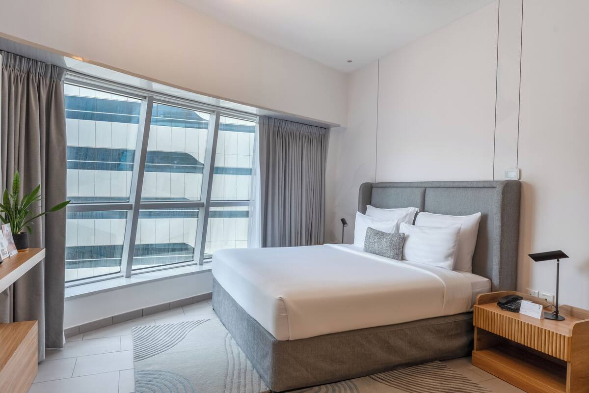 A modern hotel room with a large bed, a desk with a telephone, a small side table, a plant, and a large window with curtains.