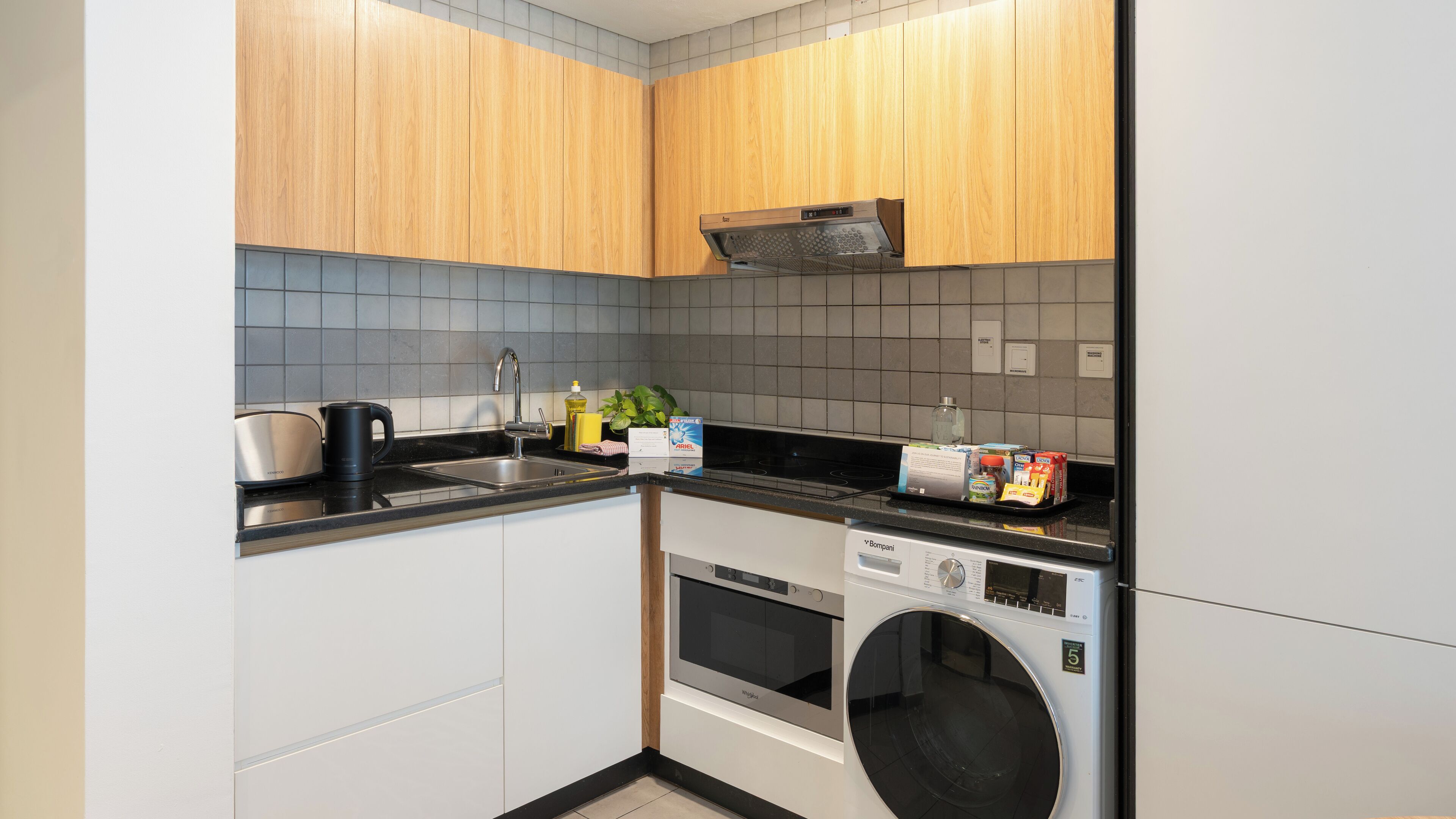 A compact kitchen with wooden cabinets, a stainless steel sink, a built-in oven, and a front-loading washing machine.