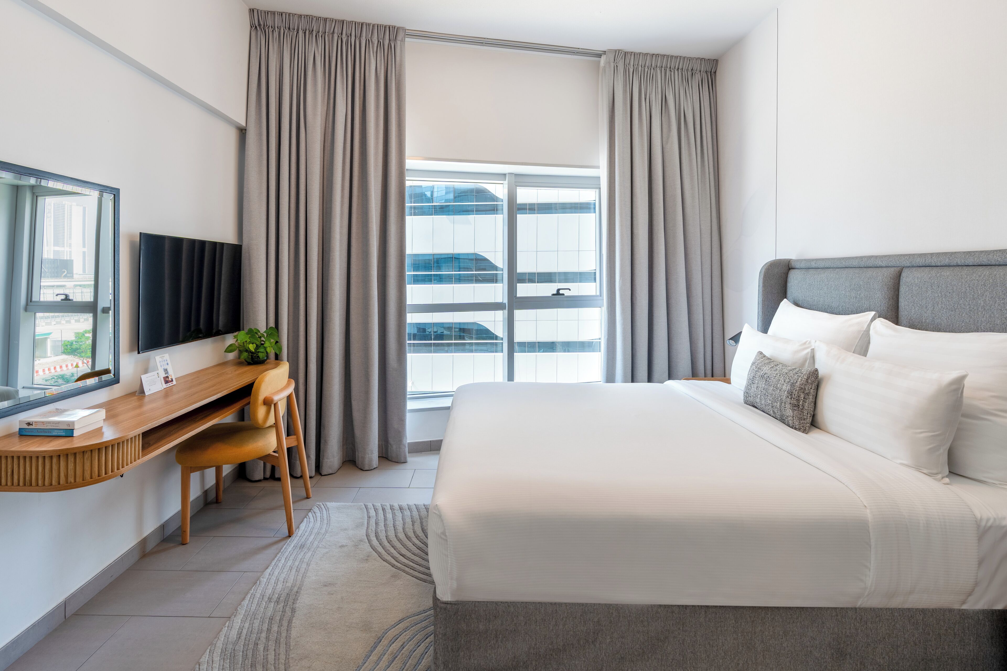 A modern hotel room with a large bed, a desk with a chair, a TV, and a window with curtains.