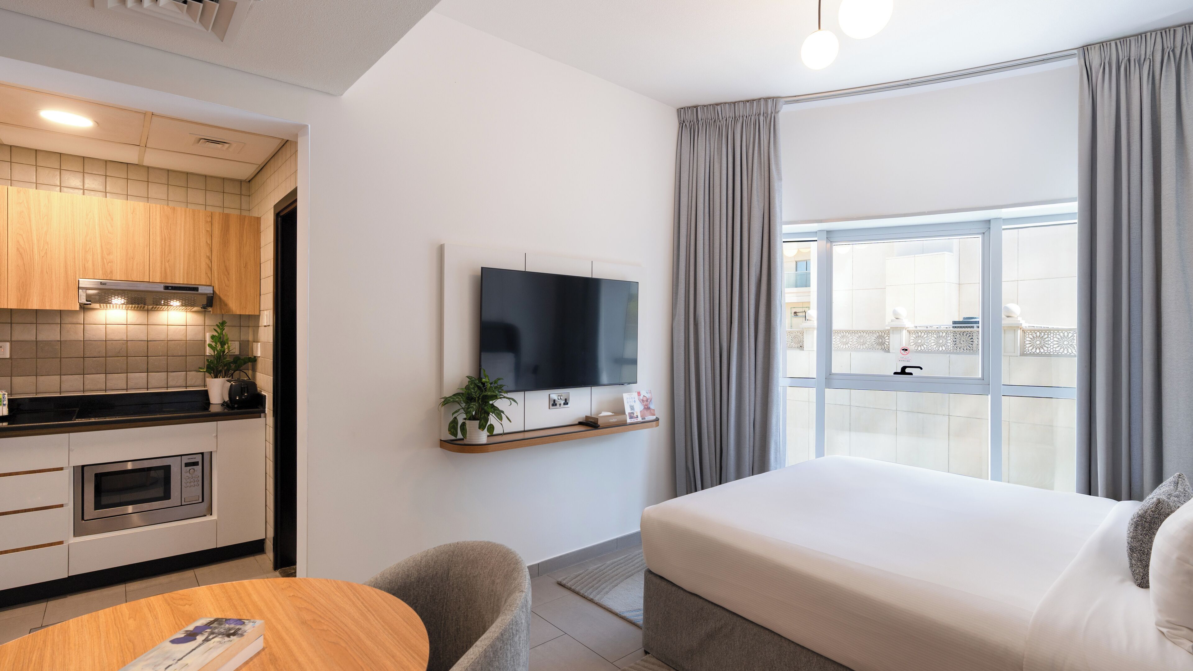 Moderni hotellihuone, jossa on sänky, seinälle asennettu televisio, mikroaaltouunilla varustettu keittiö sekä parveke, jossa on verhot.