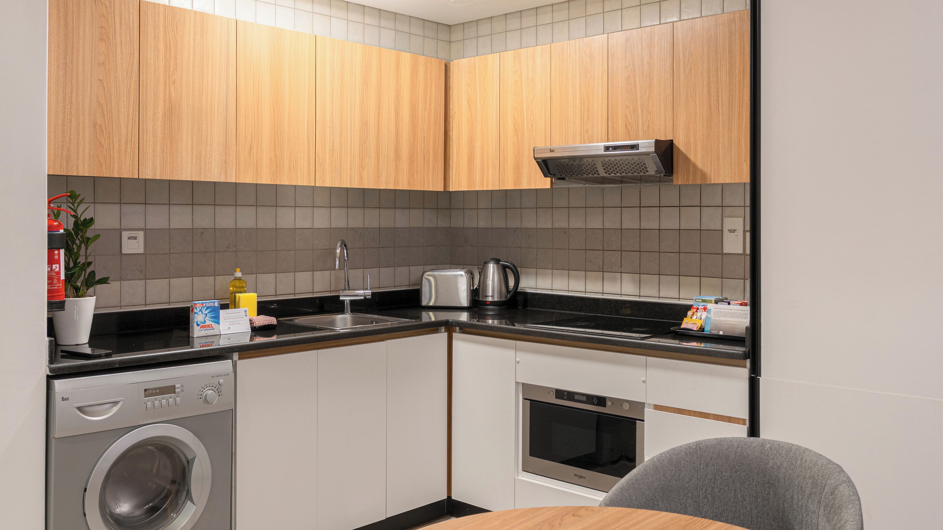 Cozinha compacta com armários de madeira, forno embutido, máquina de lavar e um pequeno espaço para refeições.