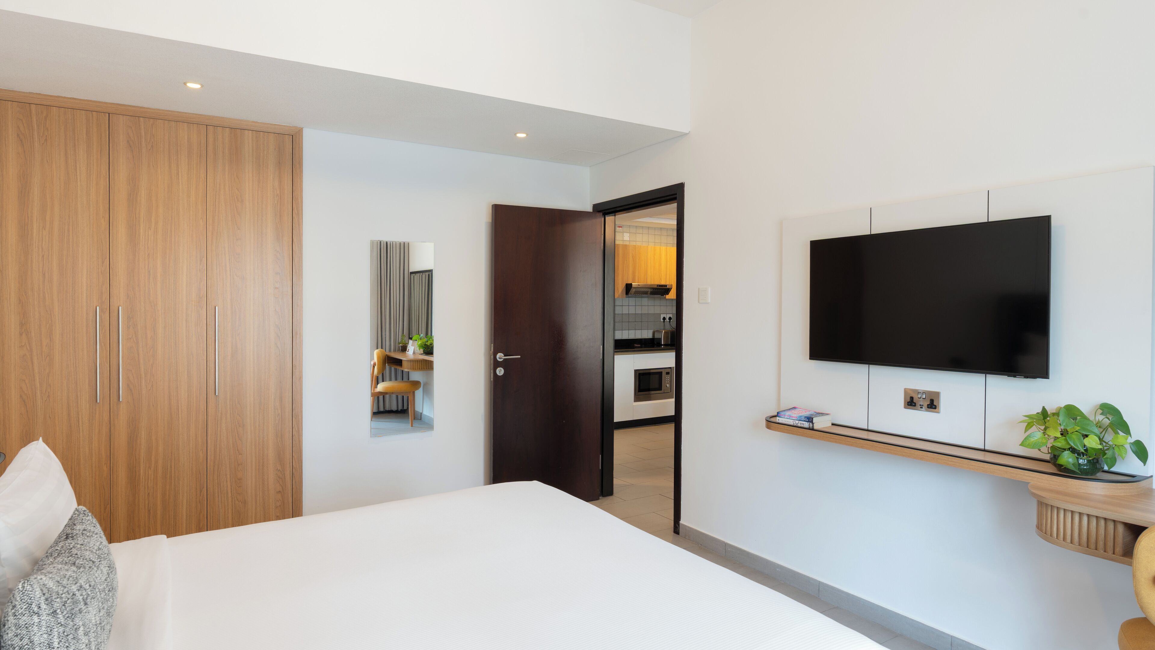 Une chambre d’hôtel moderne avec un grand lit, un téléviseur fixé au mur, une armoire en bois et un petit coin cuisine visible à travers une porte ouverte.
