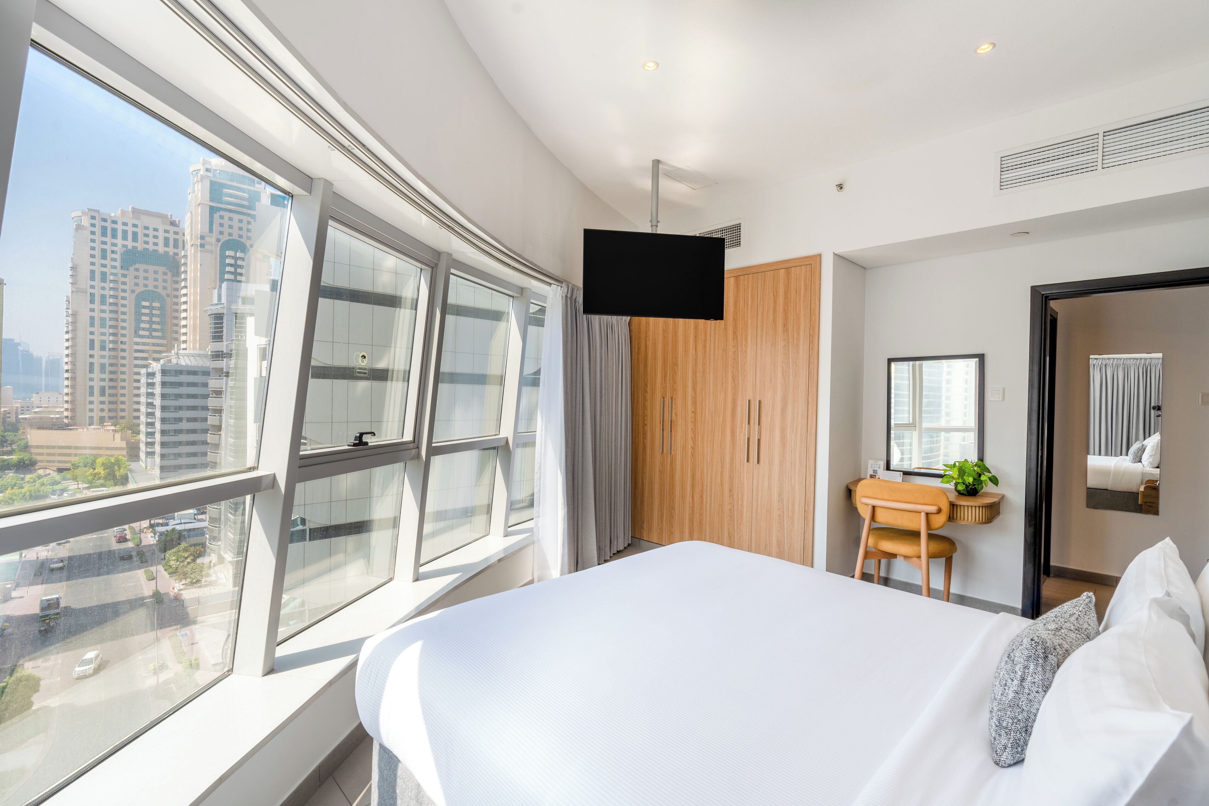 A modern hotel room with a large bed, a desk, a chair, a TV, and a view of the city through large windows.