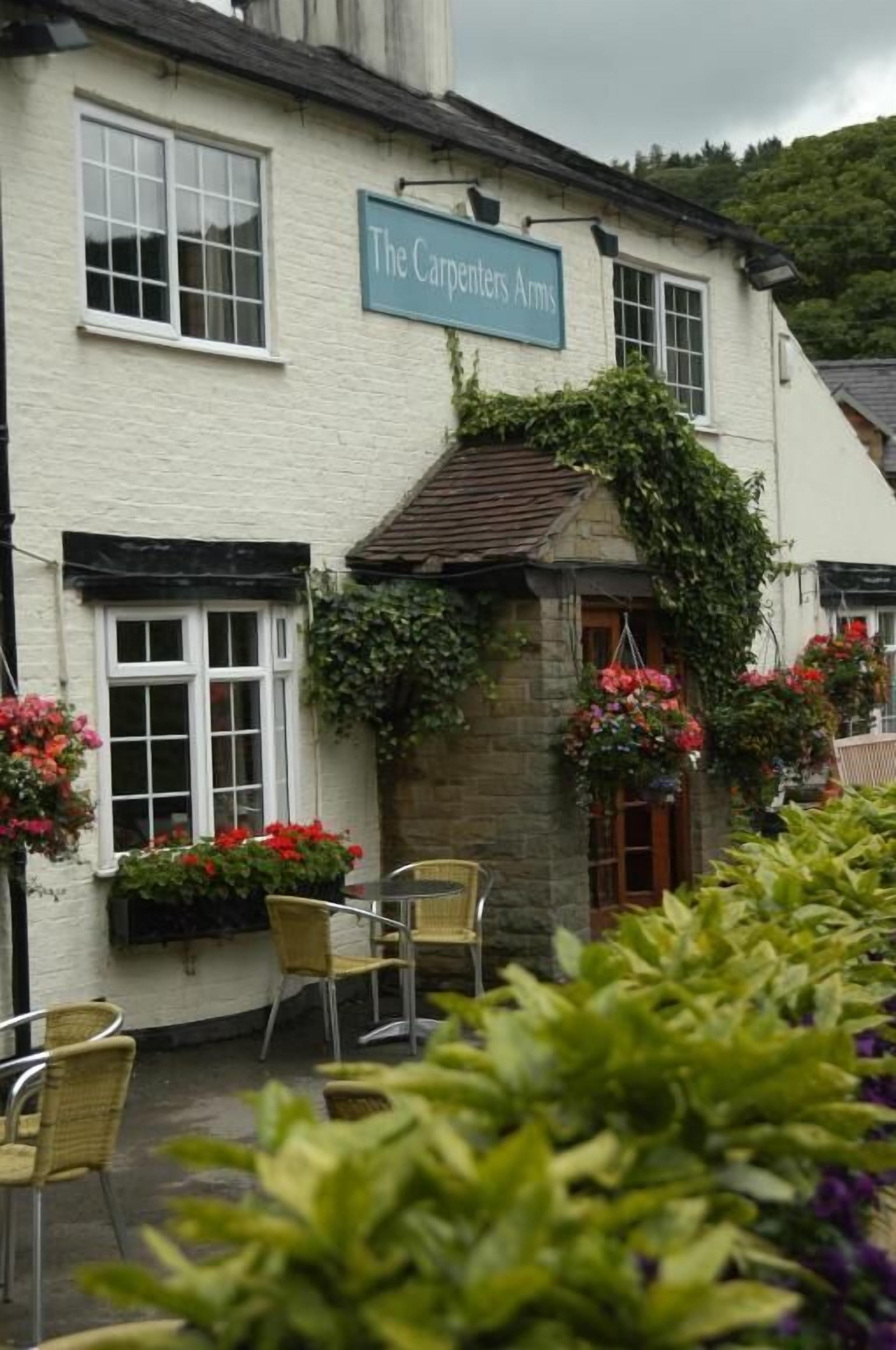 The Carpenters Arms, a white building with a sign above the entrance, outdoor seating, and hanging flower baskets.