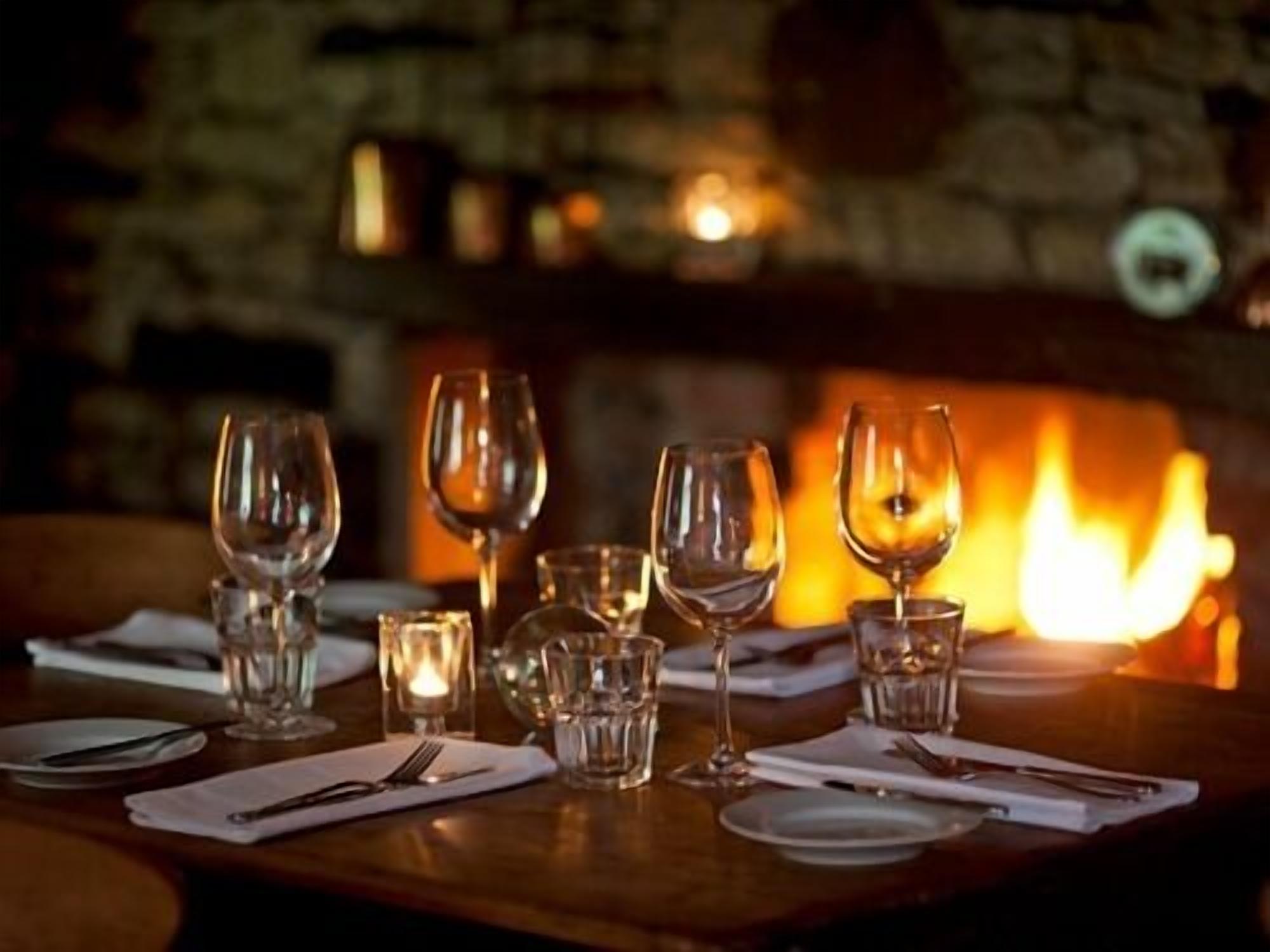 A table set for dining with wine glasses and plates, a candle, and a fireplace in the background.
