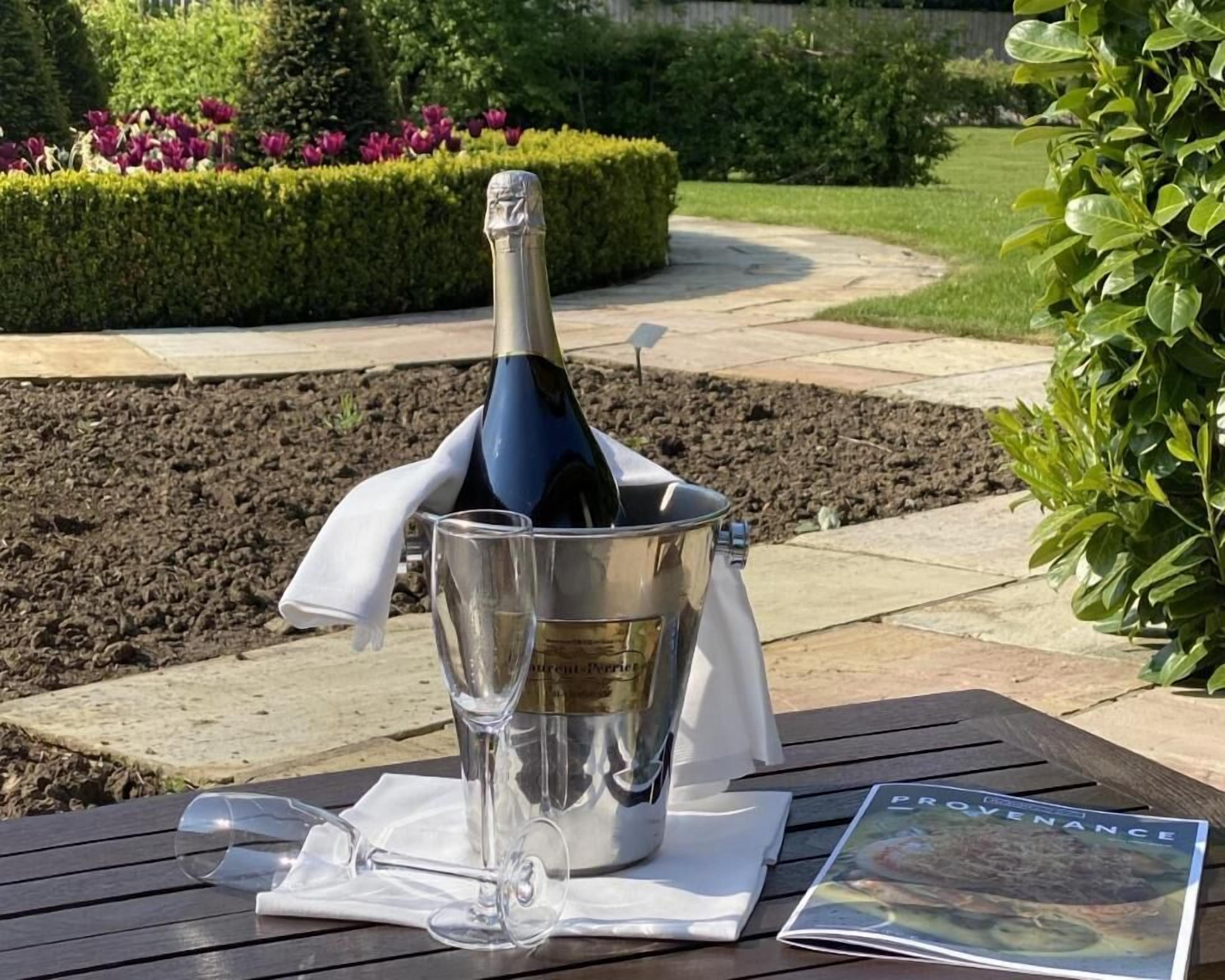 Une bouteille de champagne dans un seau en métal, posée sur une table en bois à l’extérieur.