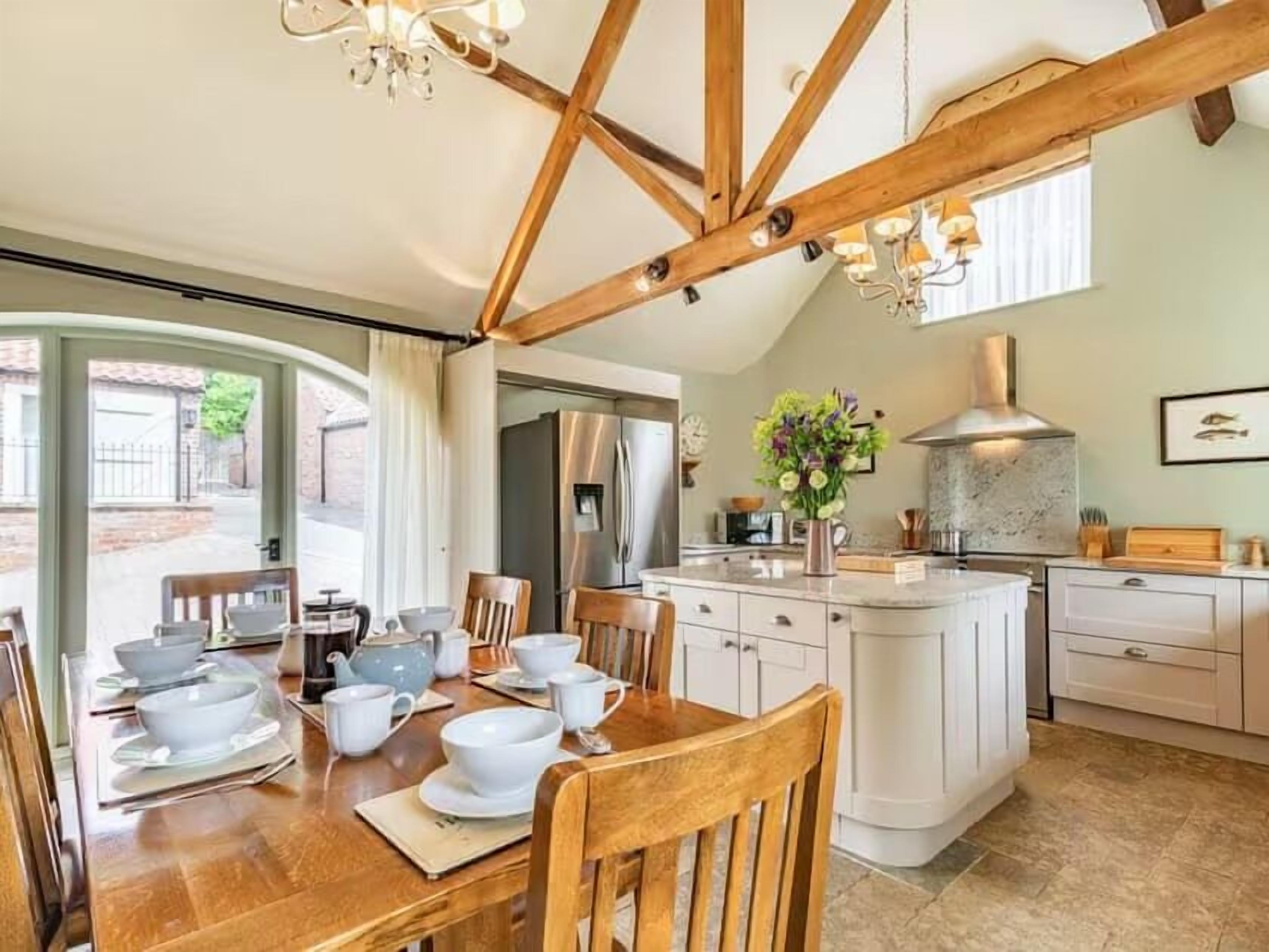 A dining area with a wooden table set for four, a kitchen with a central island, and a view through a glass door to the outside.