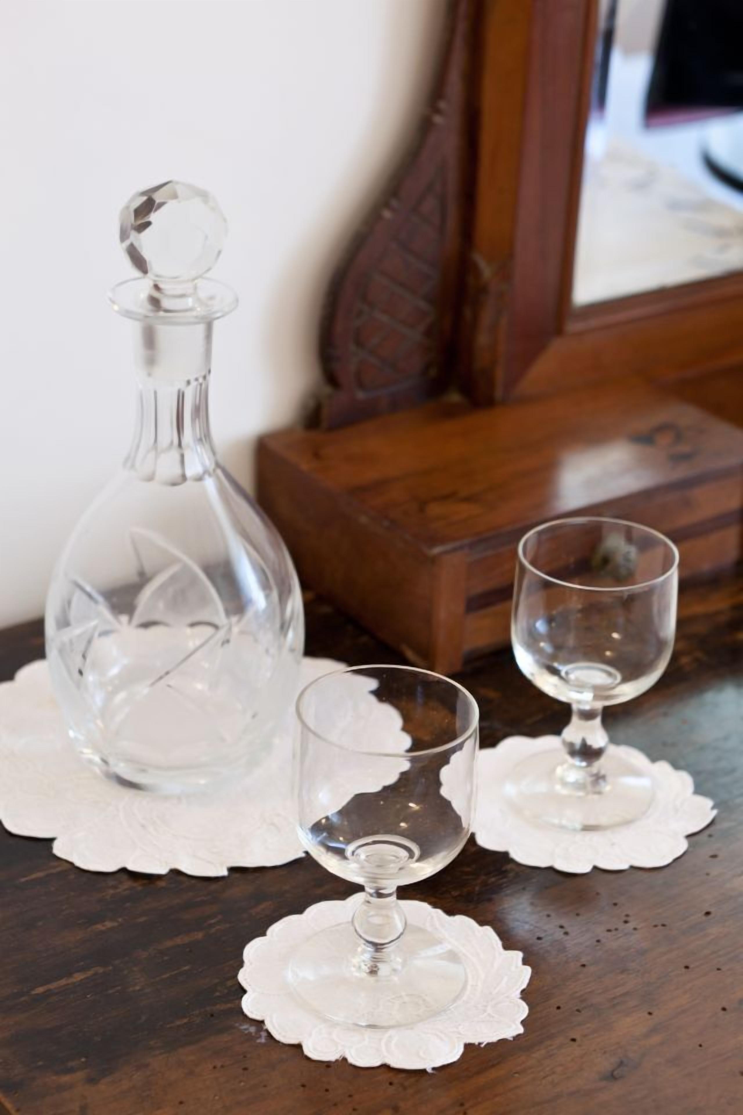 A clear glass decanter and two wine glasses on a wooden table with a lace doily.
