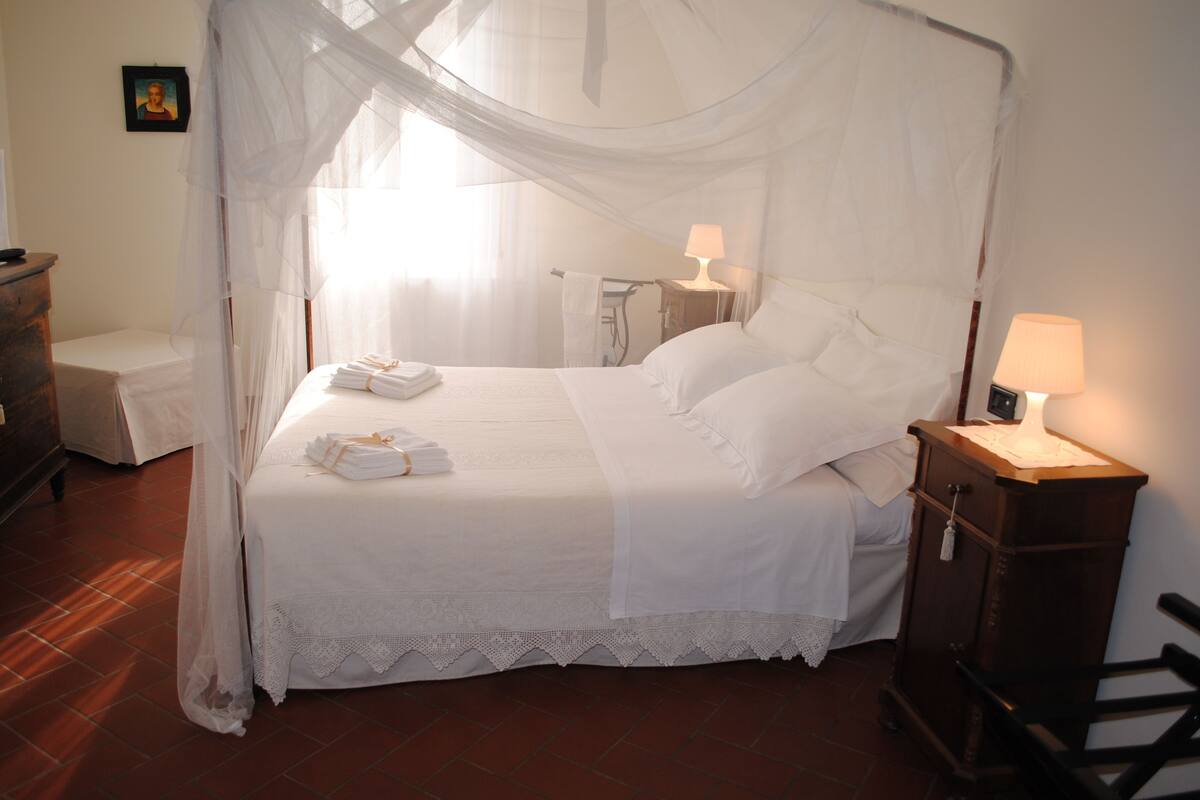 A neatly made bed with a canopy, a bedside table with a lamp, and a small wooden chair.