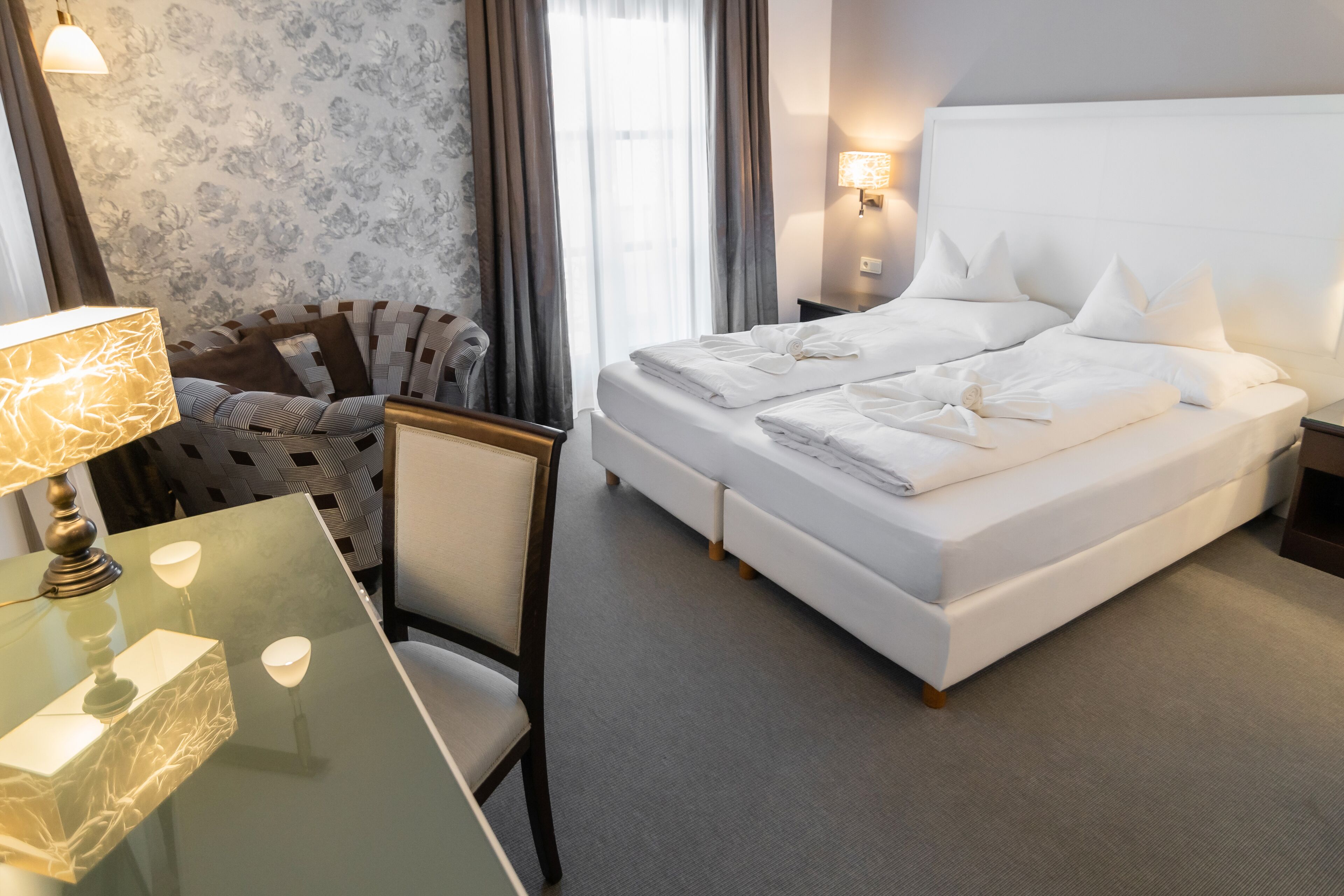 Ein Hotelzimmer mit einem großen Bett, einem Glastisch mit Lampe, einem Sessel und einem Nachttisch ebenfalls mit Lampe.