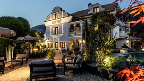 A well-lit outdoor seating area with multiple chairs and tables, surrounded by lush greenery and a pool. - Hotel Villa Groff (Ora)