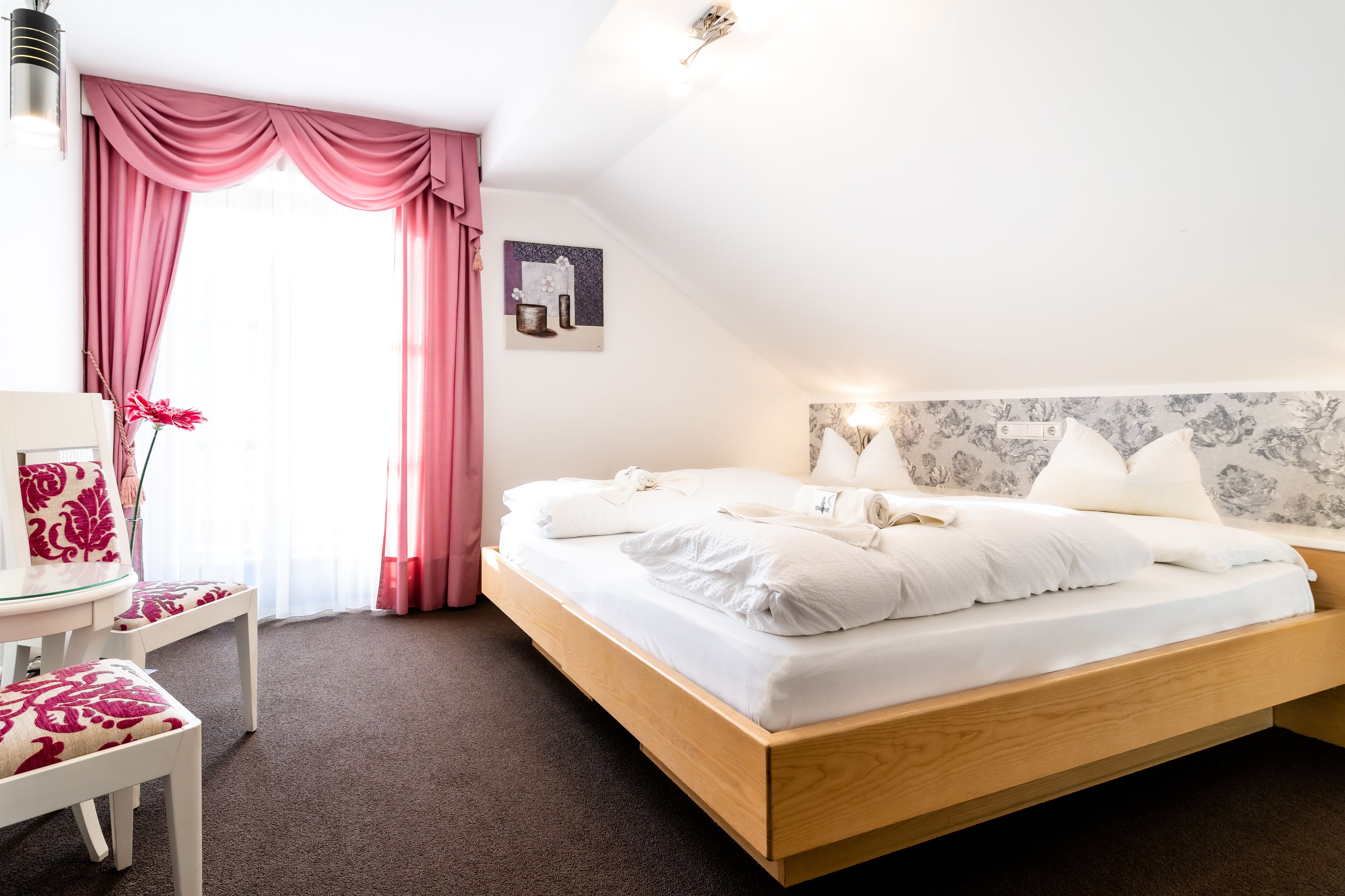 Une chambre à coucher avec un lit en bois, une literie blanche, un bureau avec une chaise et une fenêtre ornée de rideaux roses.