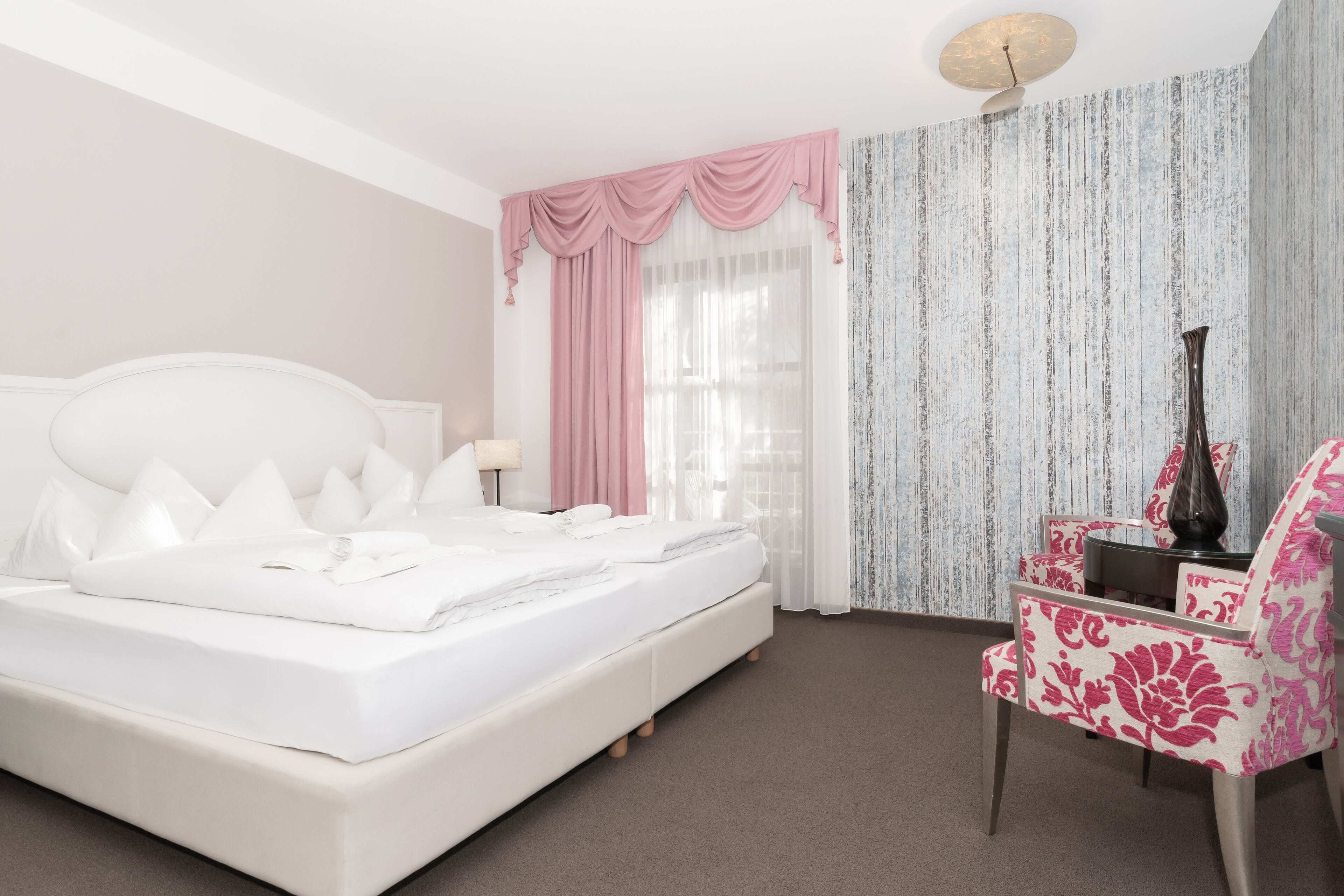 Un dormitorio con una cama grande, un escritorio con silla, un jarrón y una ventana con cortinas.