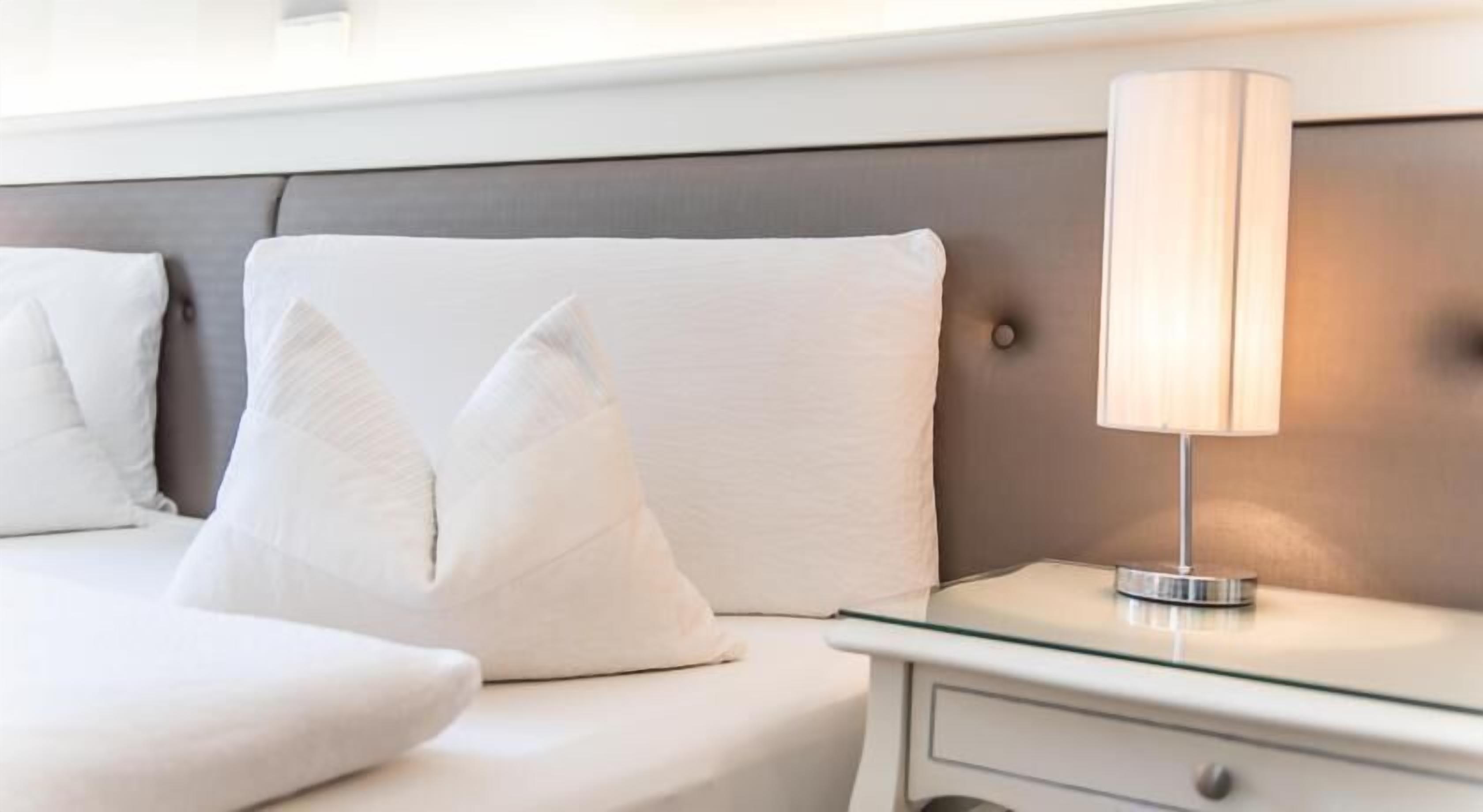 Una cama bien tendida con sábanas y almohadas blancas, una mesita de noche con lámpara y un cabecero moderno.