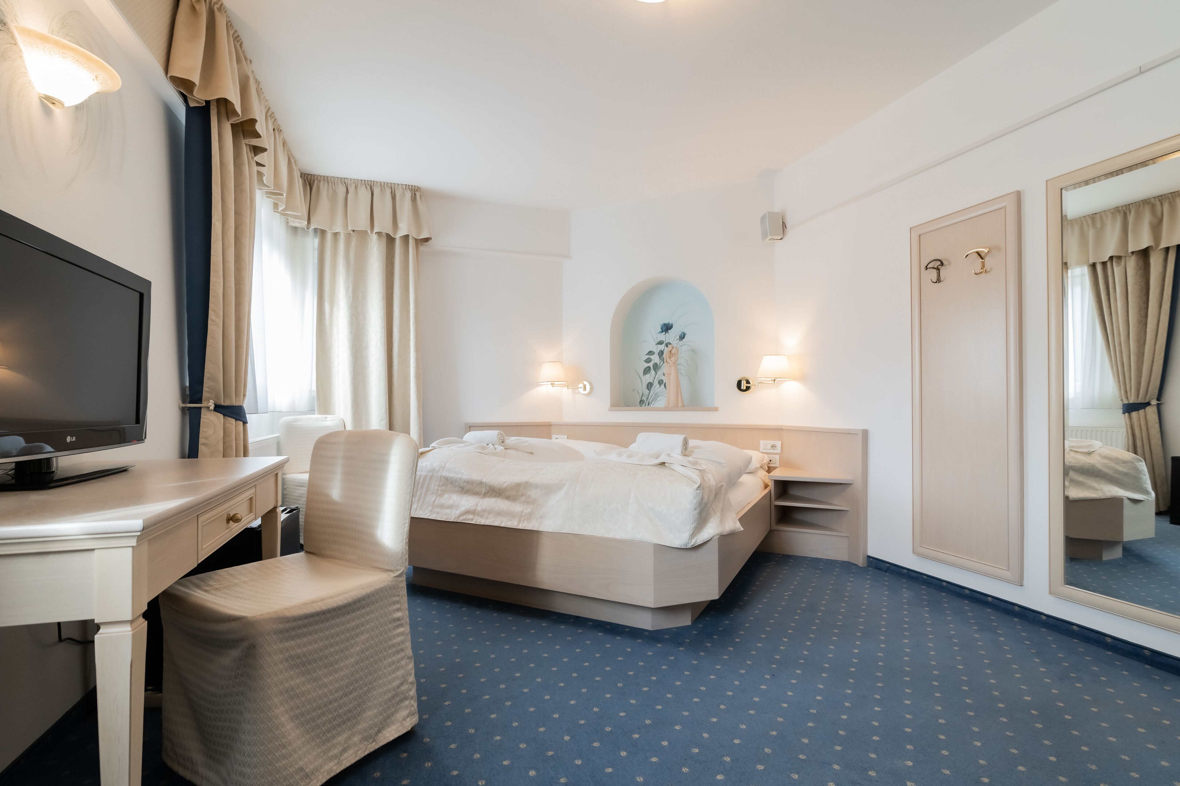 Habitación de hotel con cama, escritorio, silla, televisor, espejo y ventana con cortinas.